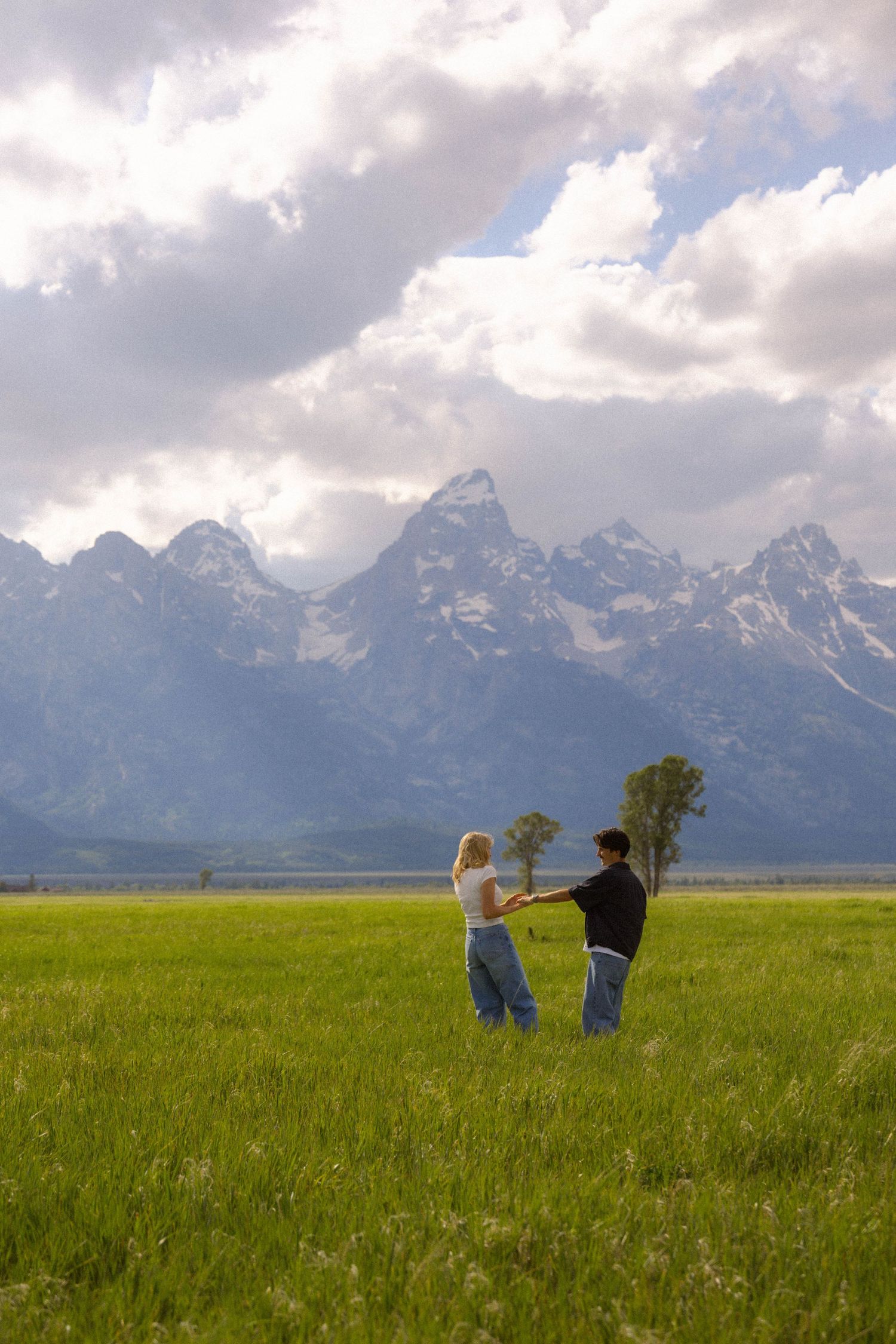 Couple holding hands dancing in green meadow with Grand Teton mountain range and dramatic clouds behind them.