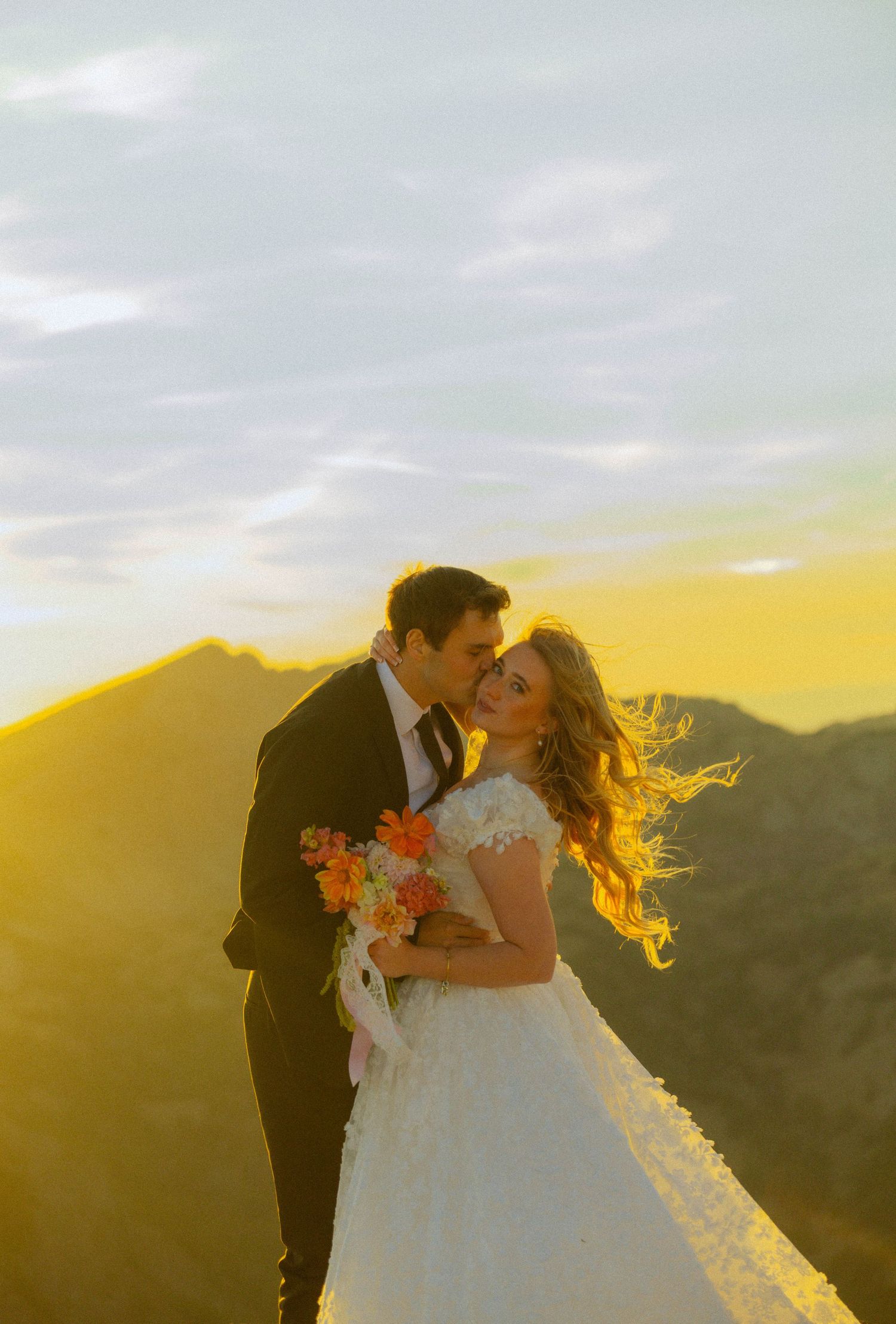 Couple in wedding attire embracing at golden sunset on mountain peak, bride holding orange bouquet with hair blowing in wind.