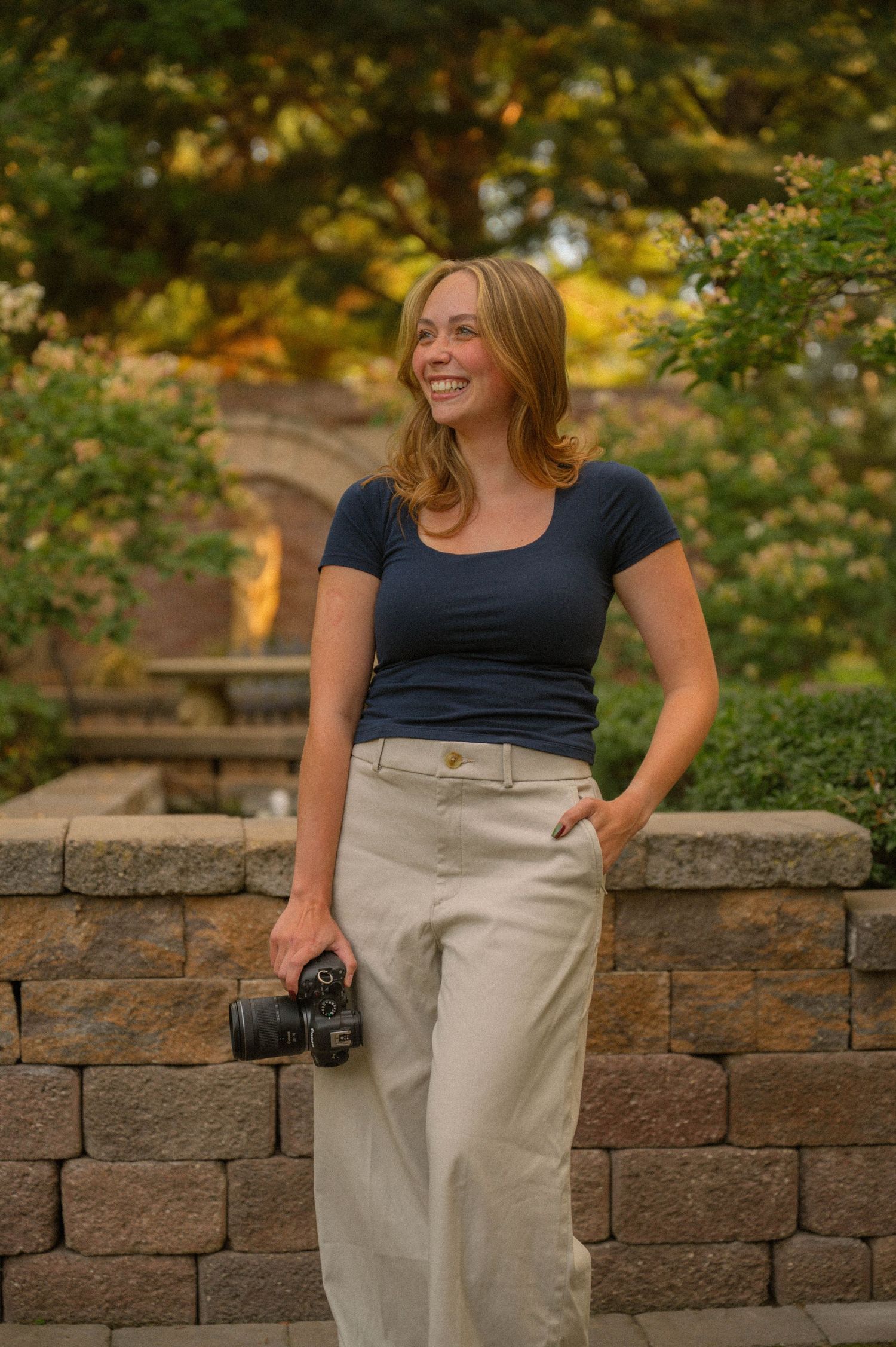 Smiling woman in navy top and khaki pants holds a camera outdoors near a stone wall with lush greenery.