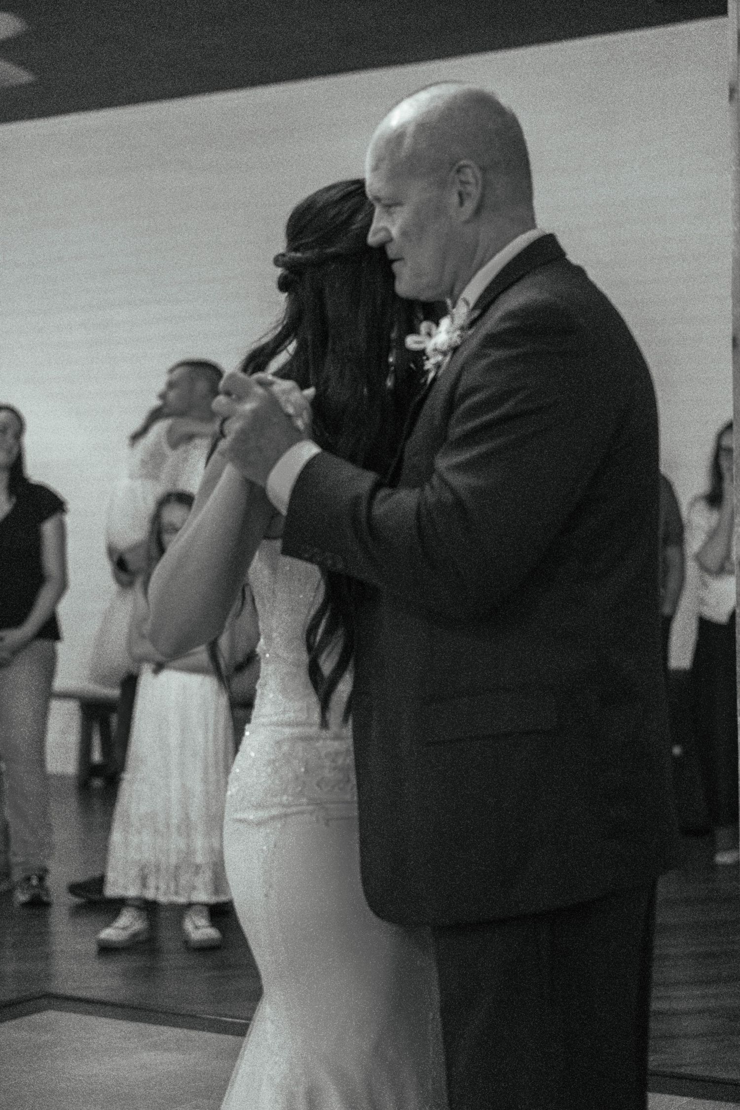 Bride in lace gown shares emotional father-daughter dance at wedding reception, black and white photo.