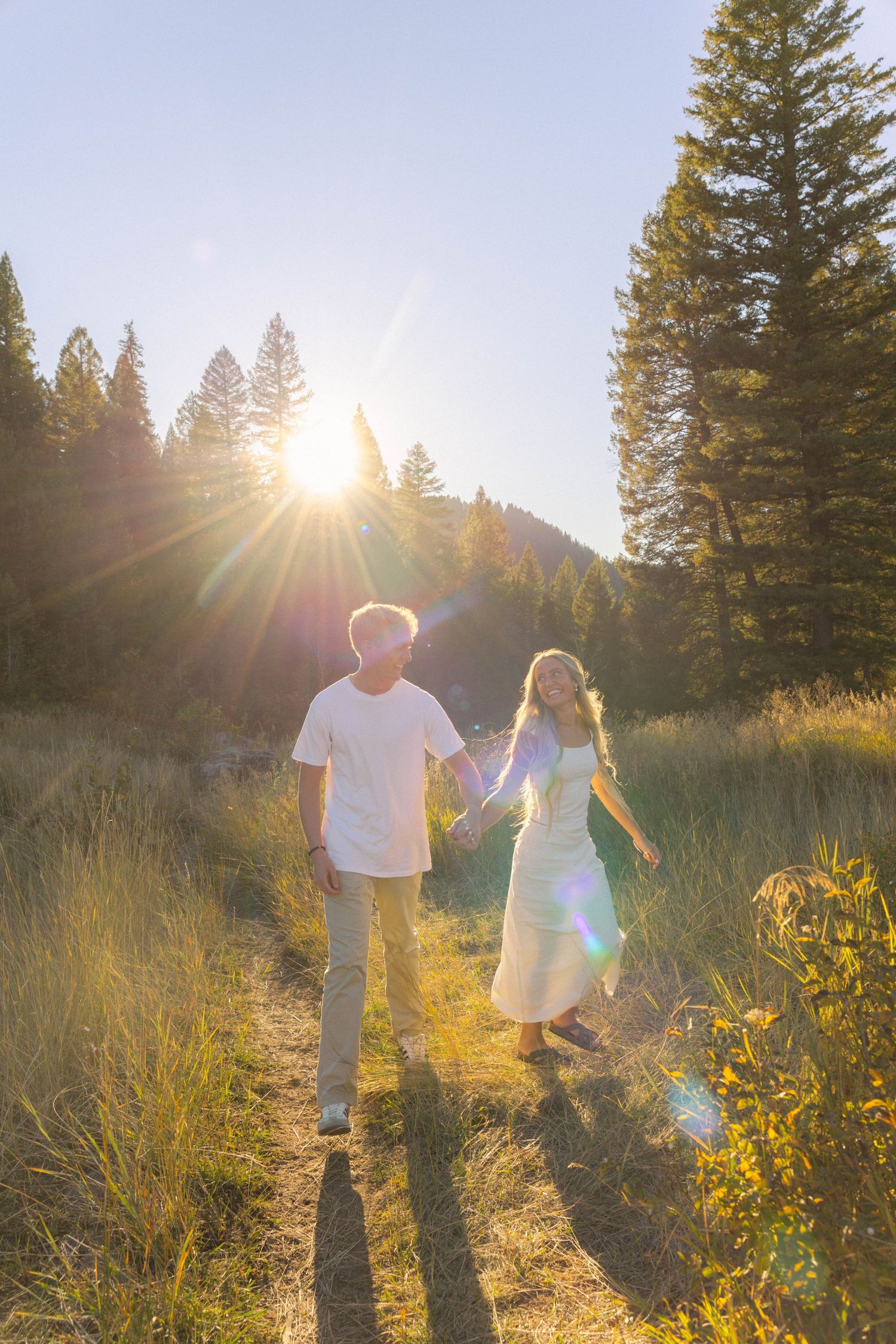 Couple holding hands walking on a sunlit forest path at golden hour, surrounded by tall pine trees.