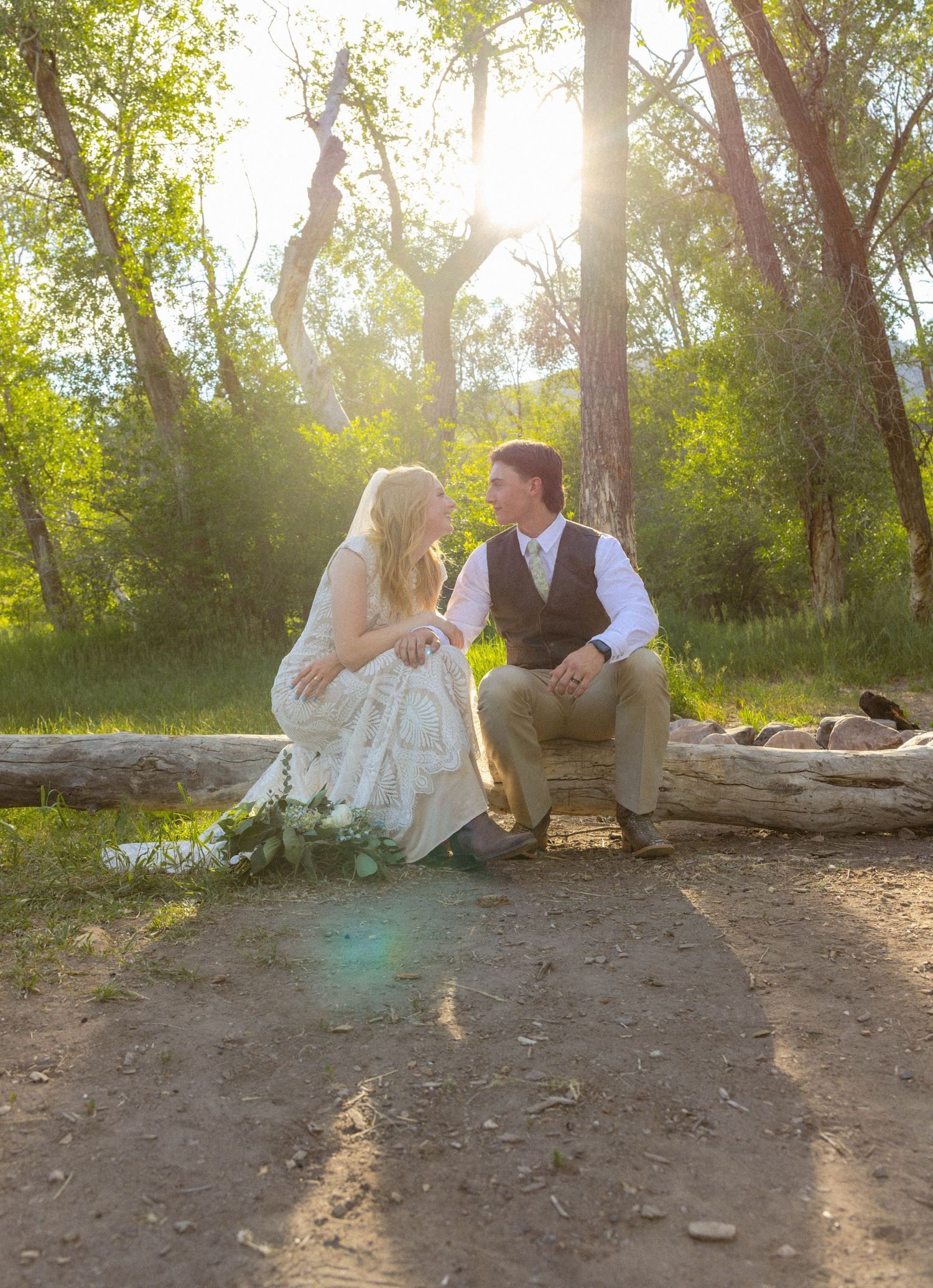 Bride and groom sit on a log in a sunlit forest, sharing a tender moment in rustic wedding attire.