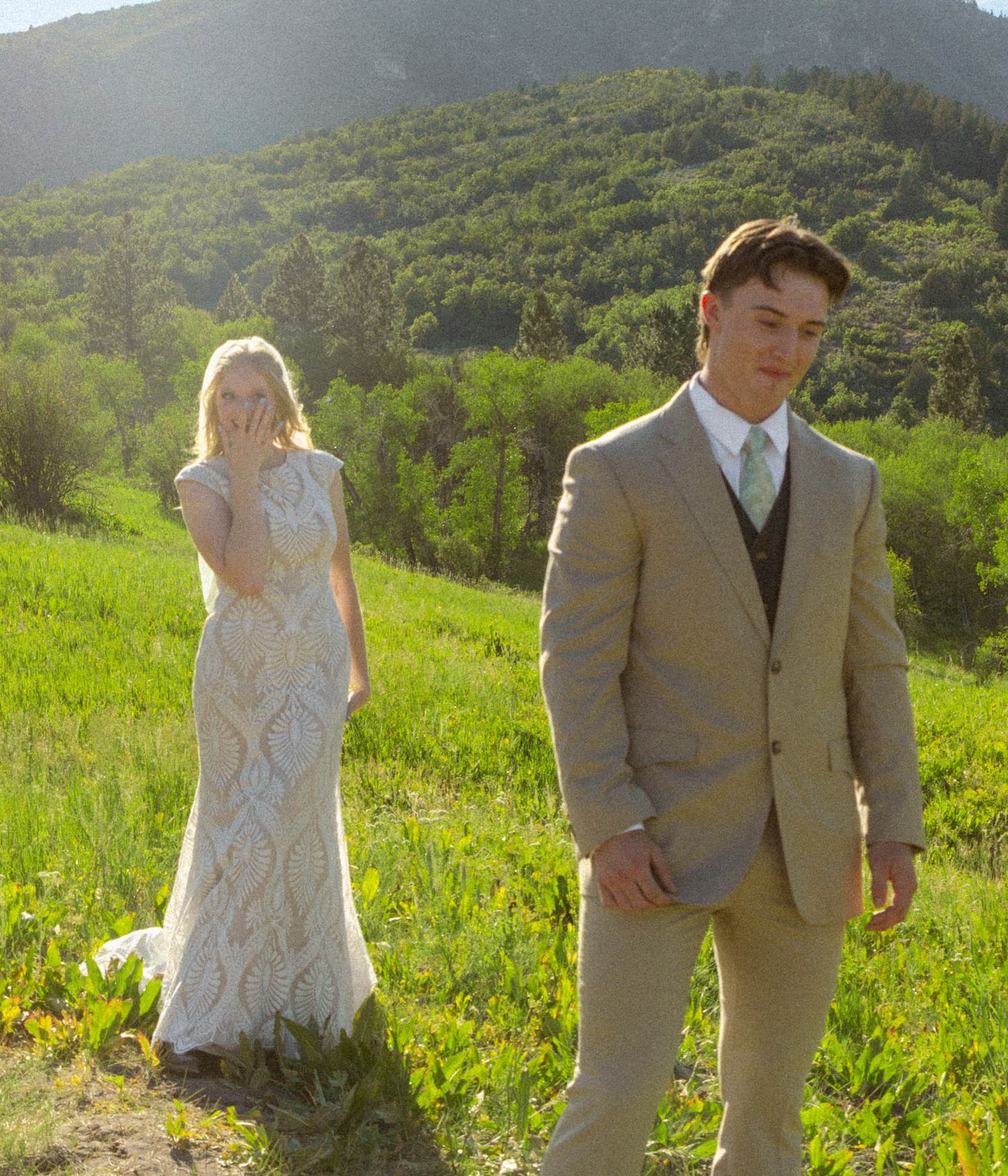 Bride in white lace gown and groom in tan suit pose outdoors on green hillside with mountain backdrop.