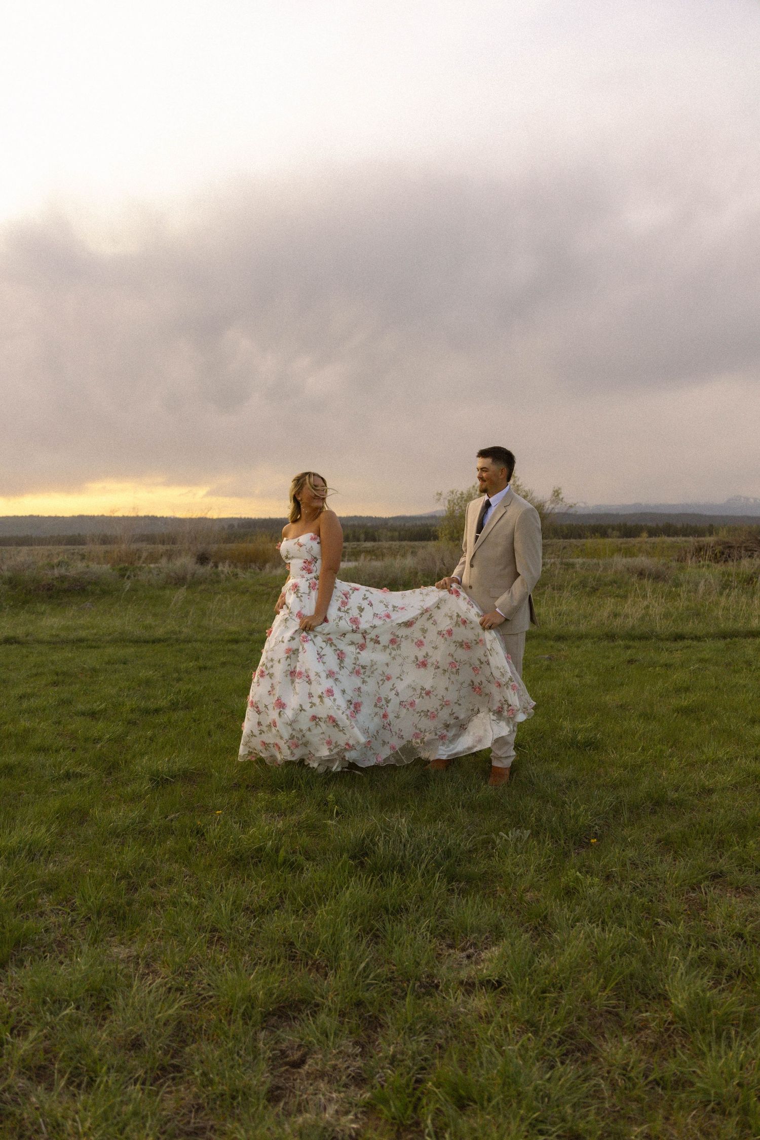 Bride in floral gown and groom in tan suit hold hands in green field at sunset with dramatic cloudy sky.