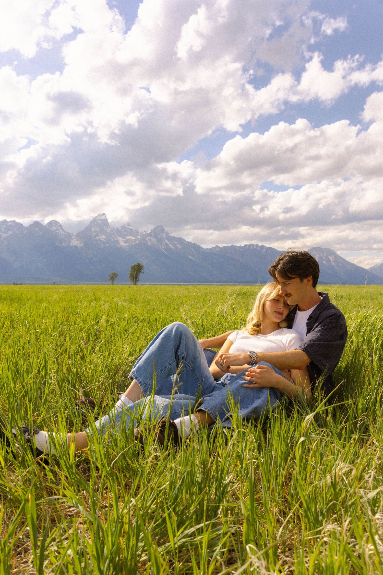 Couple sitting together in lush green meadow with mountain range and dramatic cloudy sky in background.