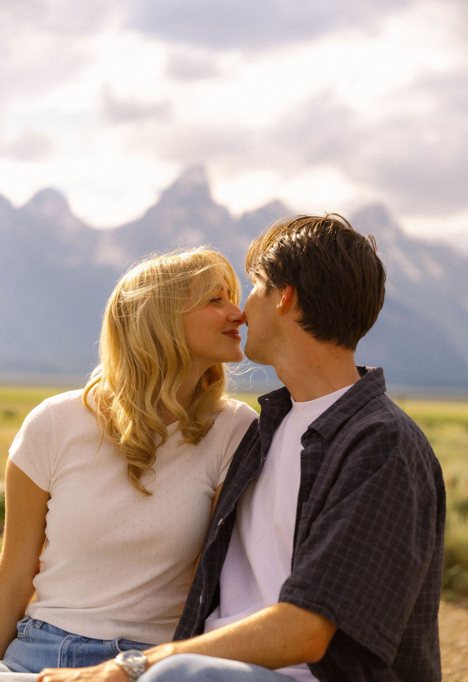 Romantic couple sharing a tender moment outdoors with majestic mountain peaks and cloudy sky in the background.