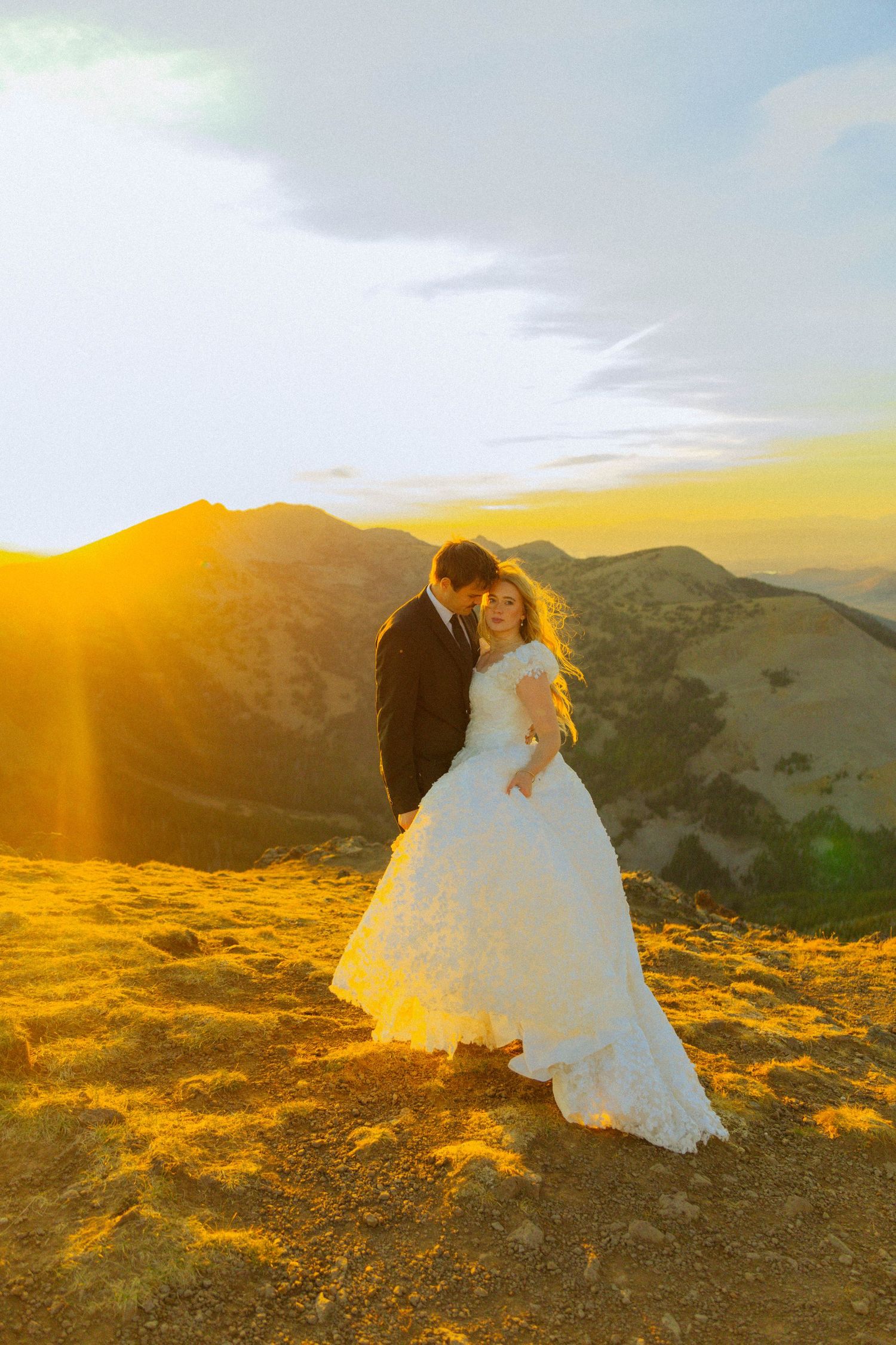 Bride and groom embracing at golden sunset on mountain peak, bride in white ball gown, romantic wedding portrait.