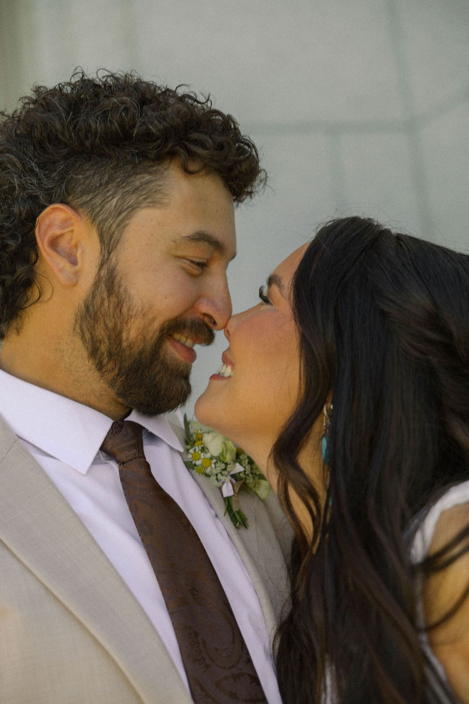 A smiling couple in wedding attire share a romantic nose-to-nose moment, groom in tan suit with brown tie and boutonniere.