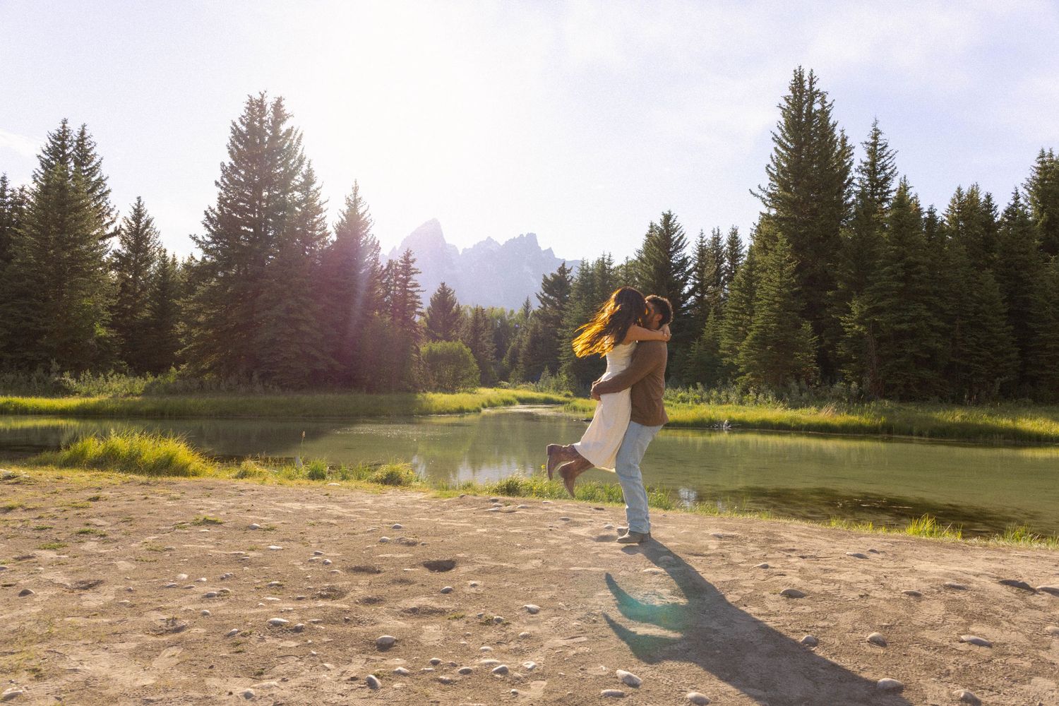 Woman spinning joyfully near a mountain lake surrounded by tall pine trees on a sunny day.