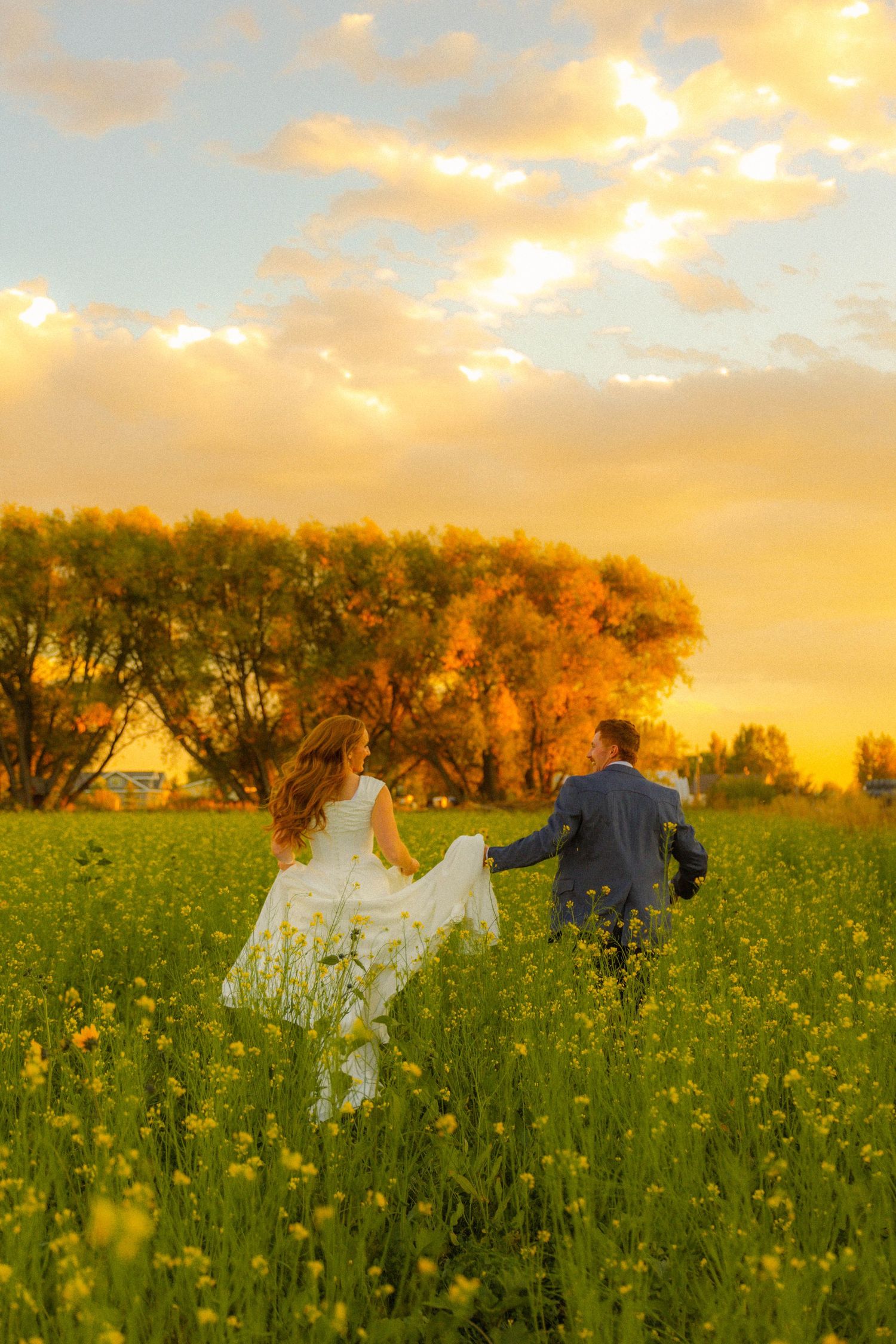 Bride and groom holding hands running through a yellow wildflower field at golden sunset with trees in background.