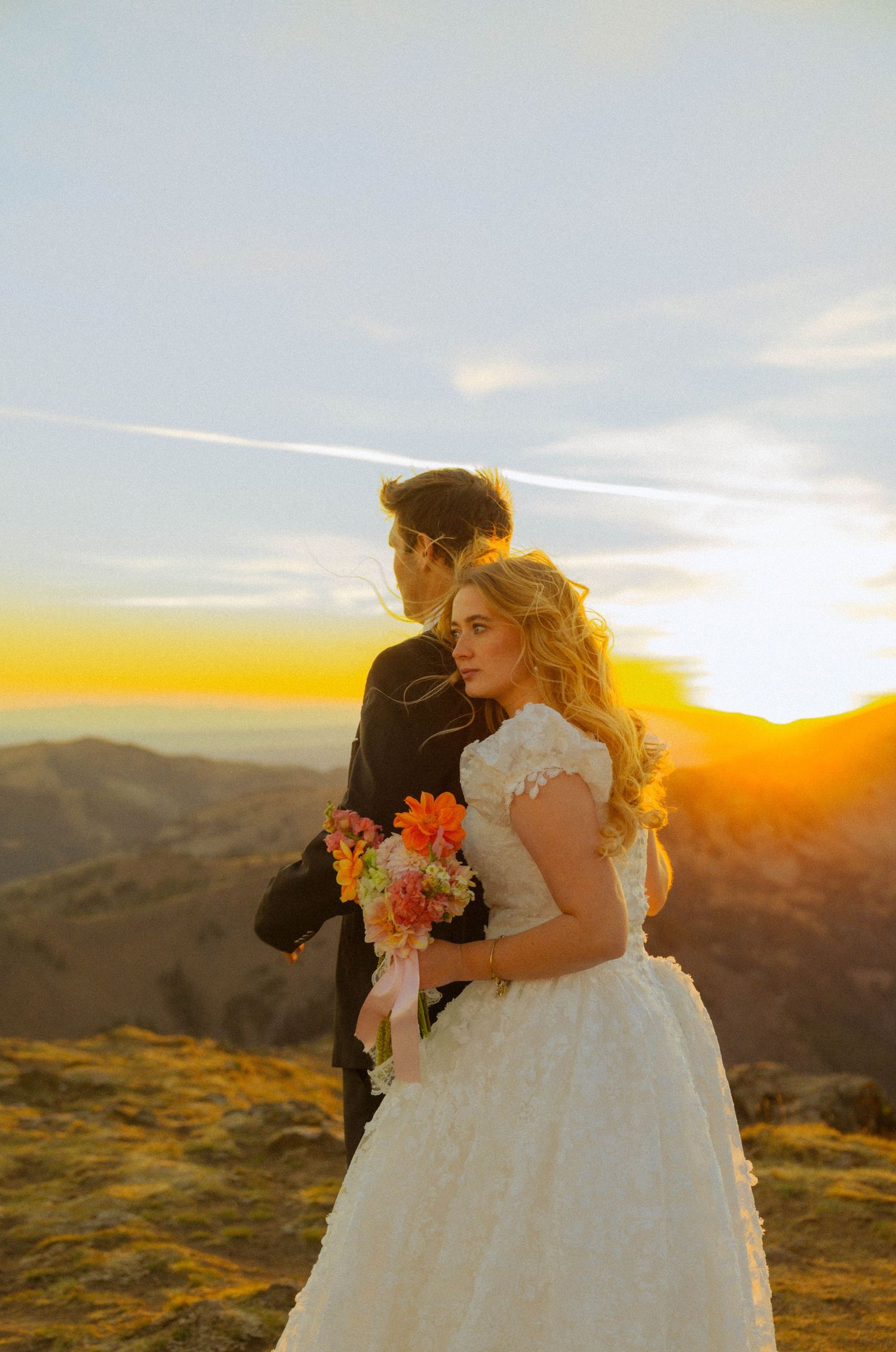 Bride in white lace gown holding colorful bouquet with groom at mountain sunset backdrop.