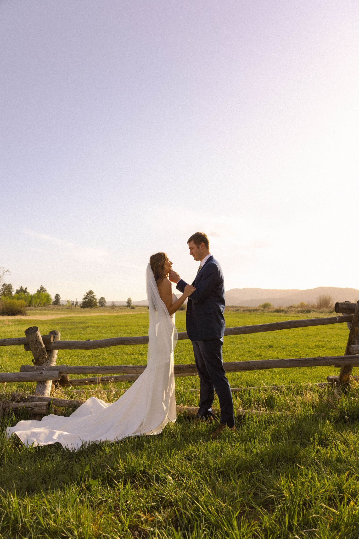 Bride and groom share a romantic moment by a rustic fence in a sunlit green meadow during golden hour.