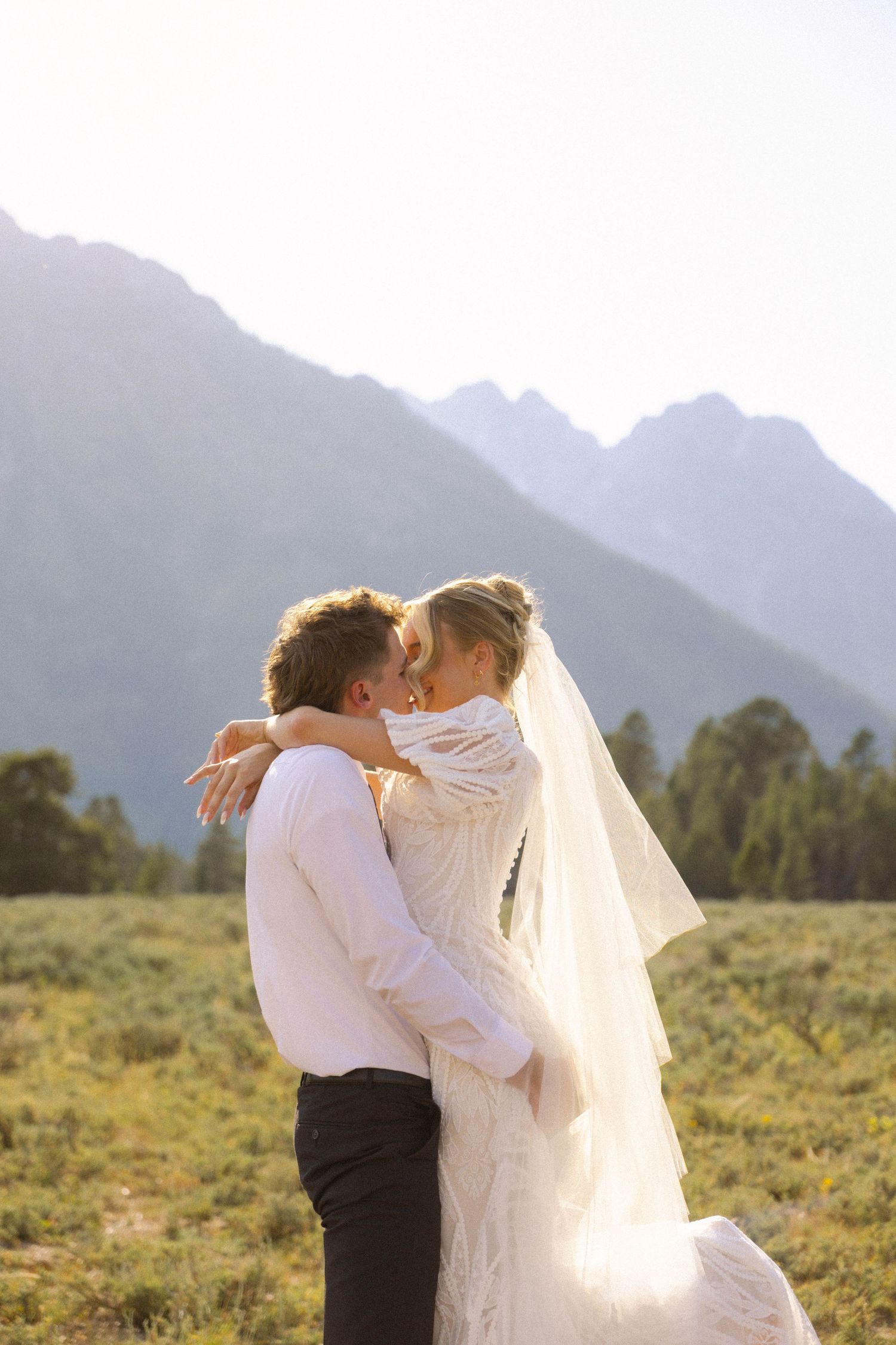 Bride and groom sharing a kiss in a scenic mountain meadow, bride wearing lace gown and veil.