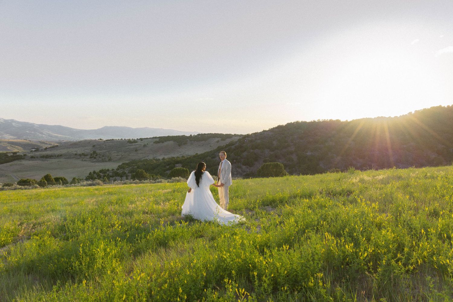 Bride and groom holding hands in a sunlit green meadow with mountain views at golden hour.