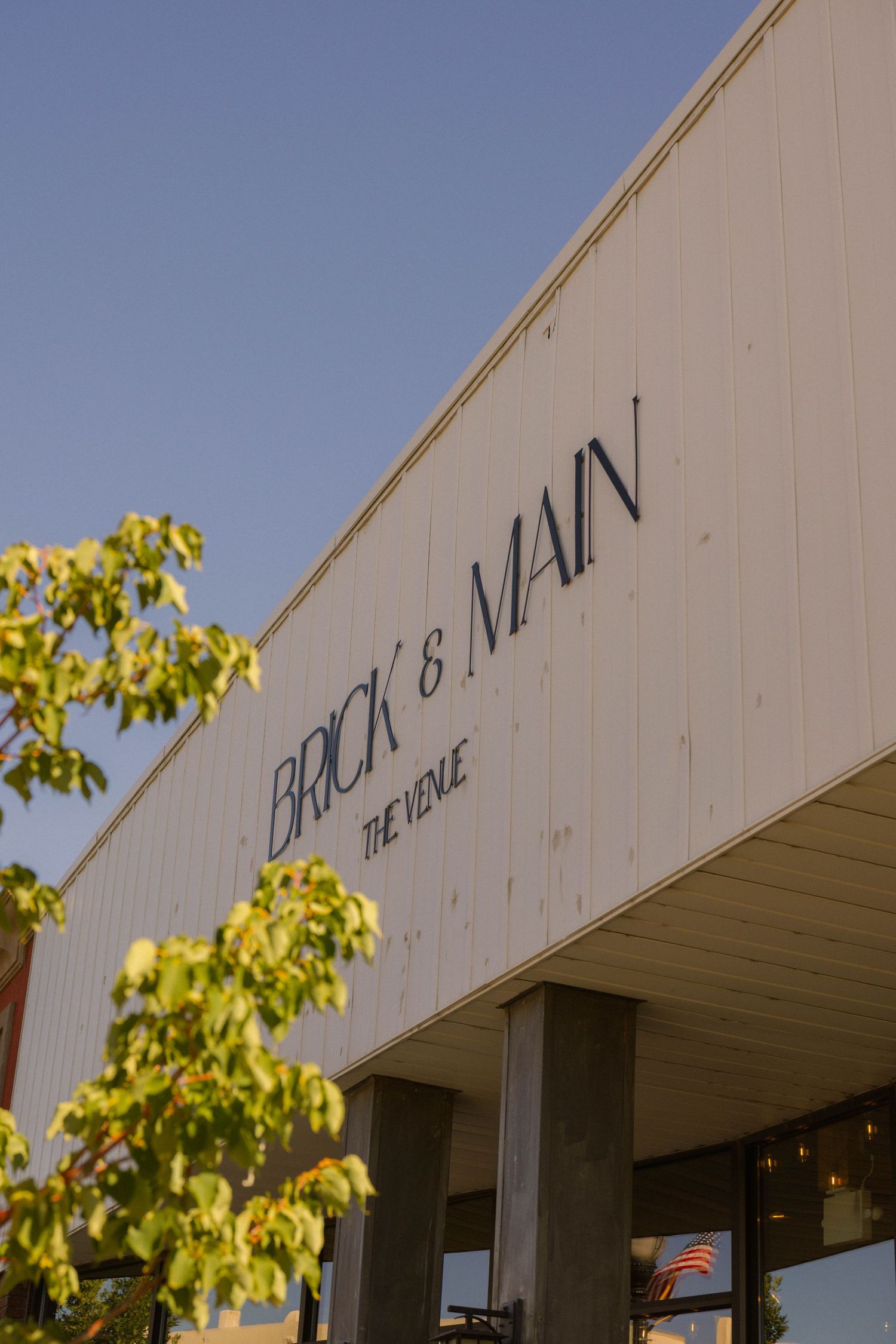 Brick & Main The Venue exterior sign on a white building facade with green tree leaves and blue sky.