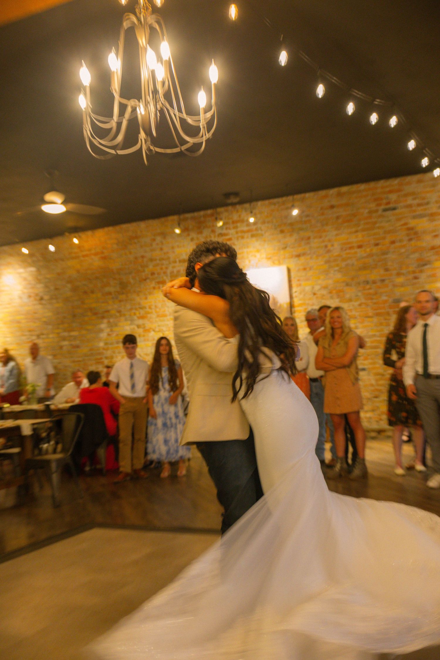 Bride and groom share their first dance under a chandelier in a rustic brick-walled wedding venue with guests watching.