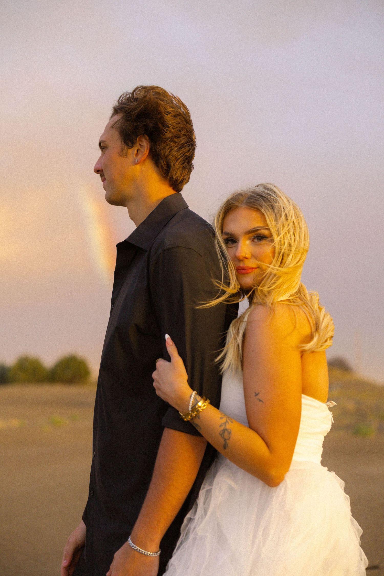Couple in wedding attire embracing at sunset with a rainbow in the background, bride in white dress looking at camera.