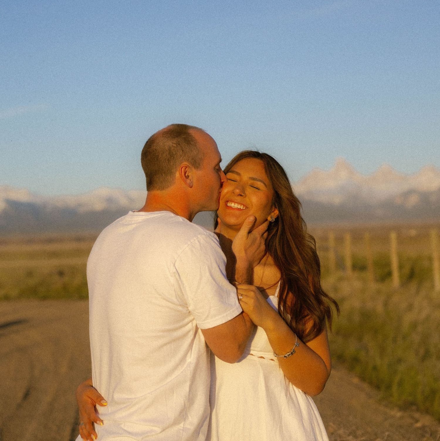 Couple sharing a romantic moment at sunset on a dirt road with mountain views, woman laughing as man kisses her cheek.