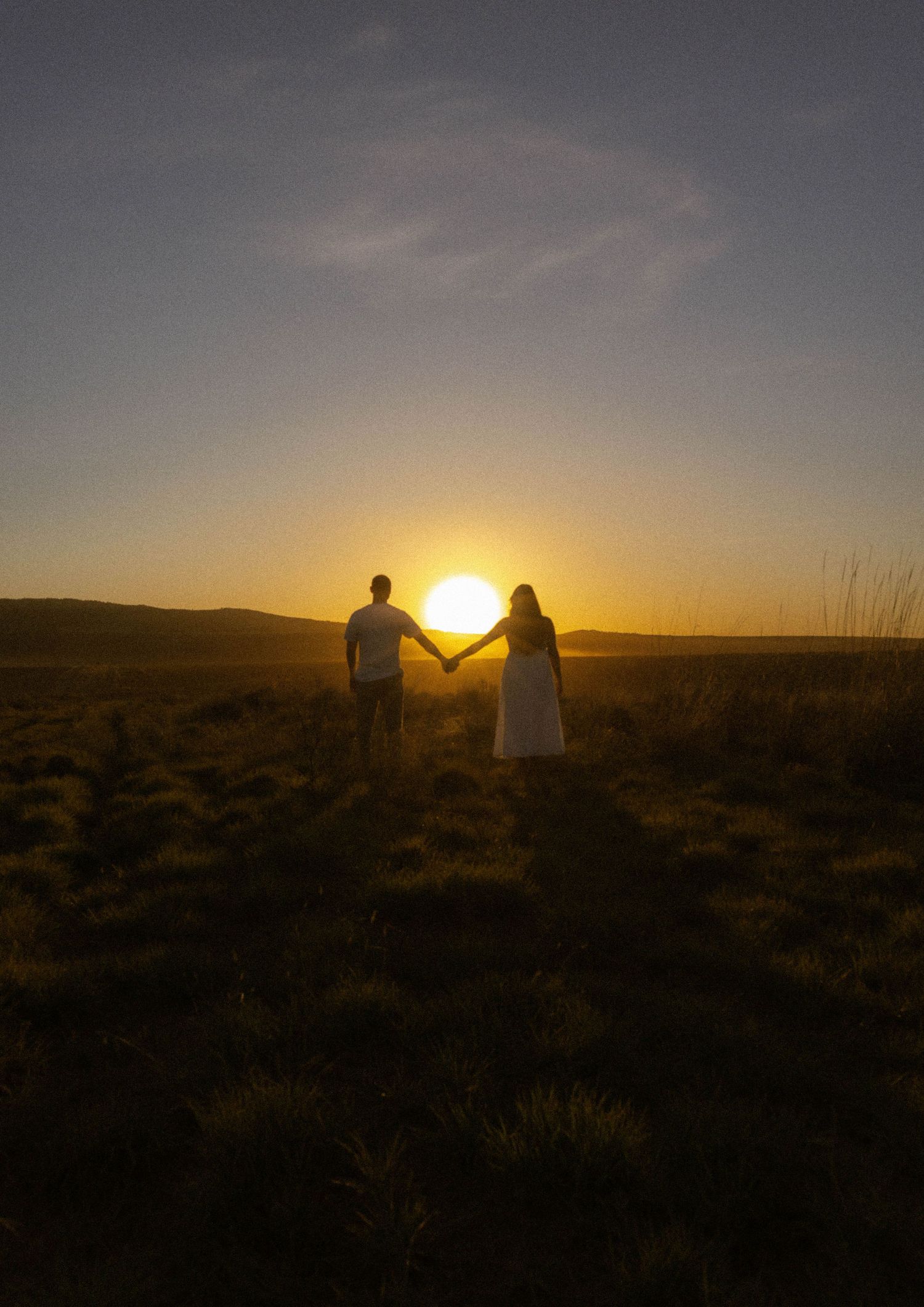 Couple holding hands silhouetted against a golden sunset in an open field.