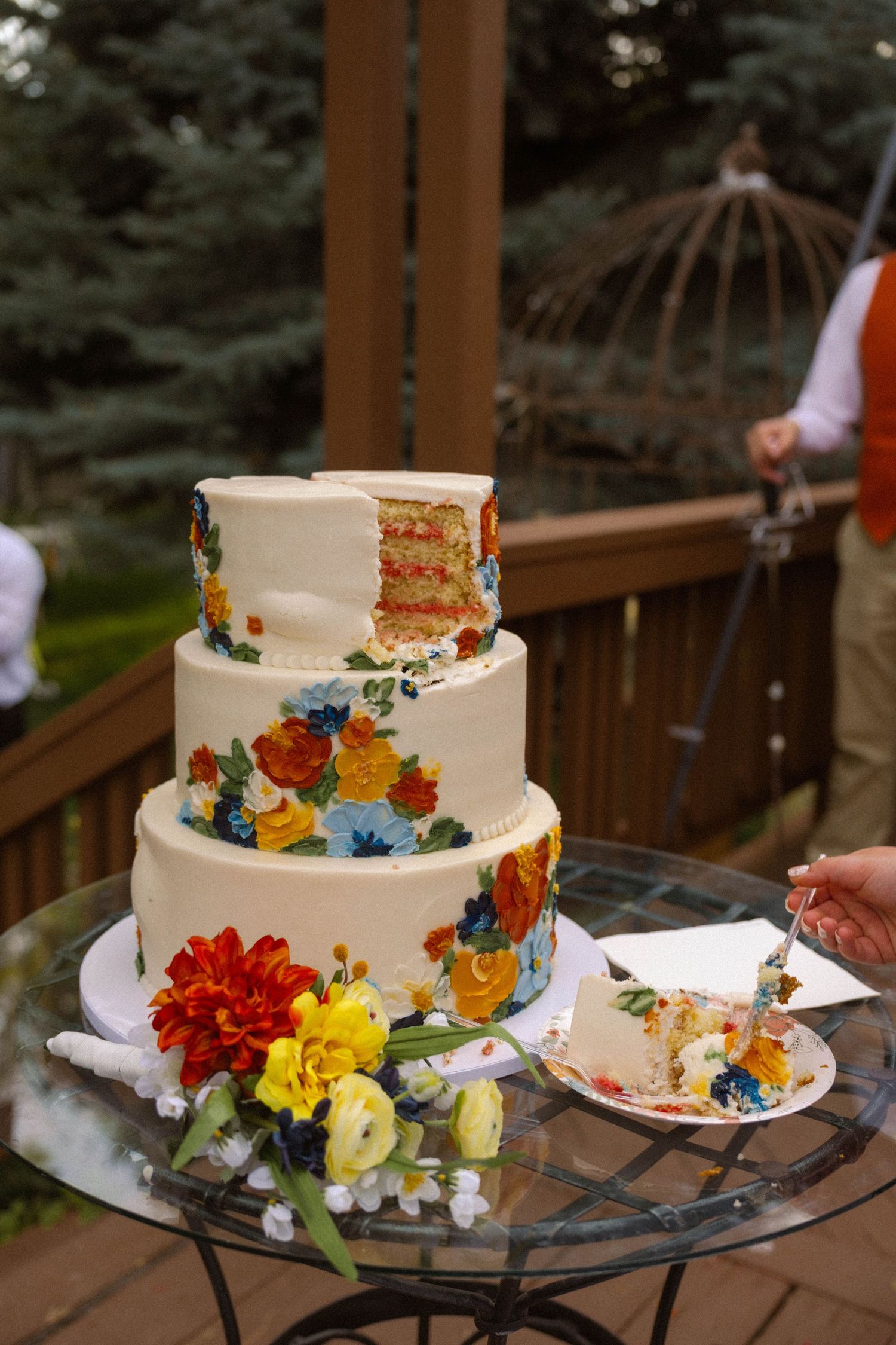 Three-tier white wedding cake with colorful floral decorations, partially sliced, displayed on glass table on wooden deck.