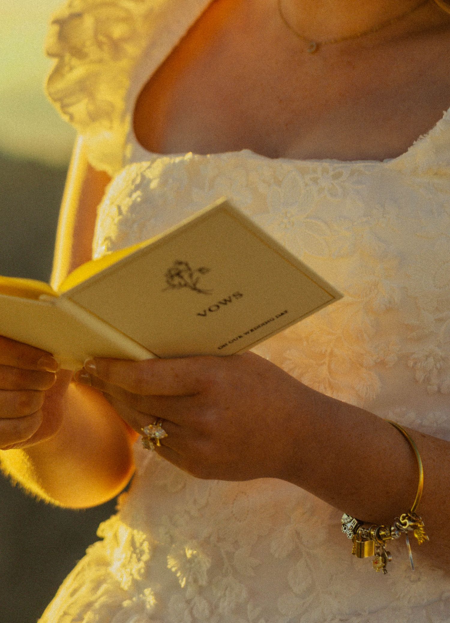 Bride in lace wedding dress reading handwritten vows from a small booklet, wearing gold bracelet and ring.