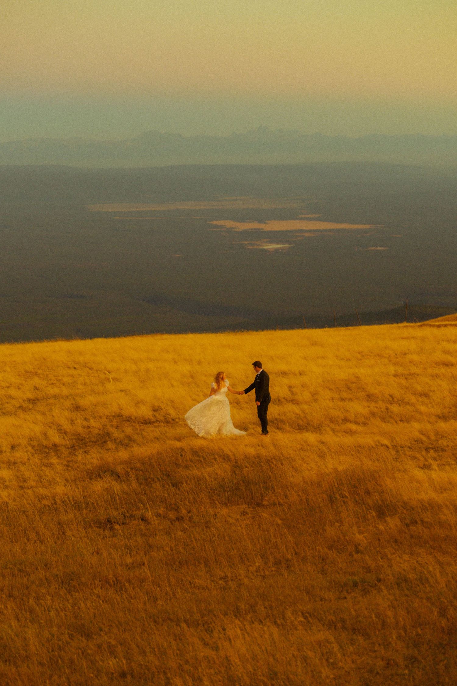 Bride and groom hold hands on golden hillside at sunset with vast mountain landscape behind them.