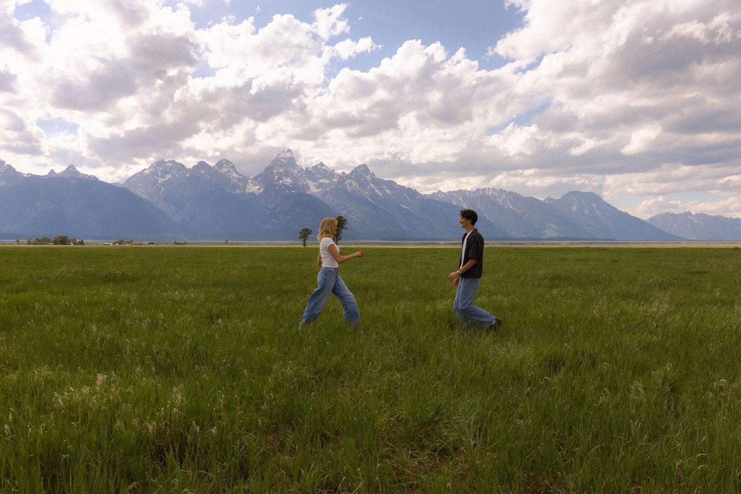 Couple walking through green meadow with Grand Teton mountain range backdrop under cloudy sky.