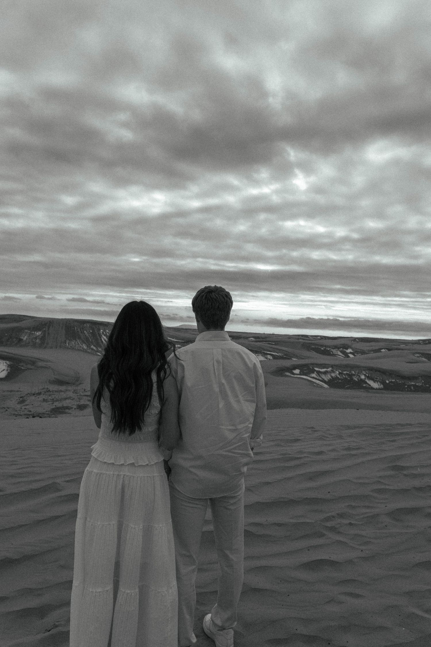 Black and white photo of a couple standing together on sand dunes under a dramatic cloudy sky.