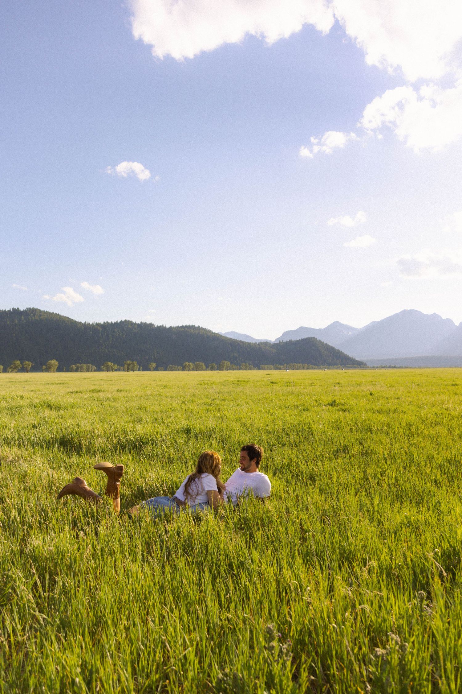 Couple sitting together in a lush green meadow with mountain views and blue sky backdrop.