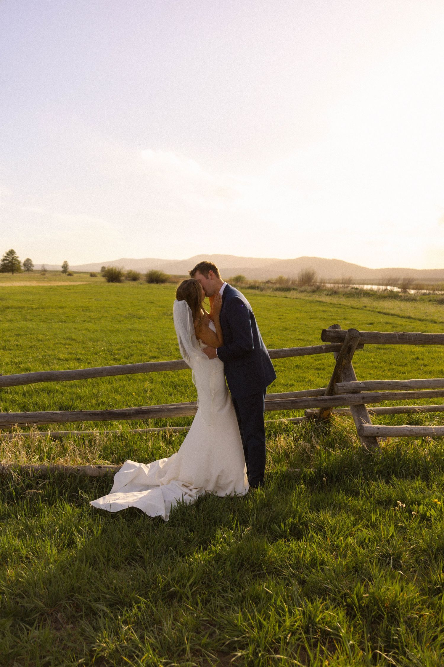 Bride and groom share a romantic kiss by a rustic wooden fence in a sunlit green meadow with mountains in the background.