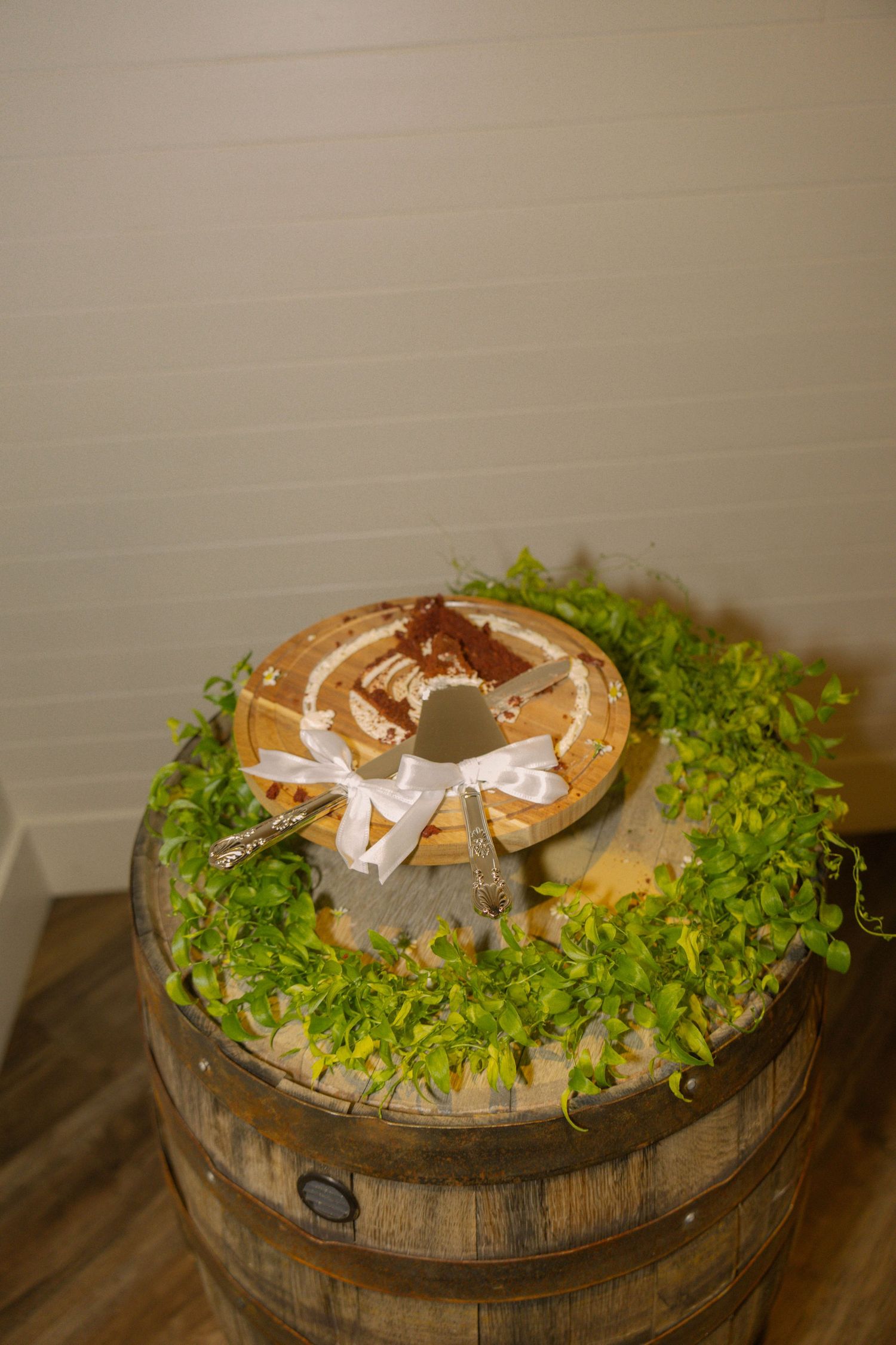 Rustic wedding cake on wooden barrel decorated with greenery and white ribbon bow.