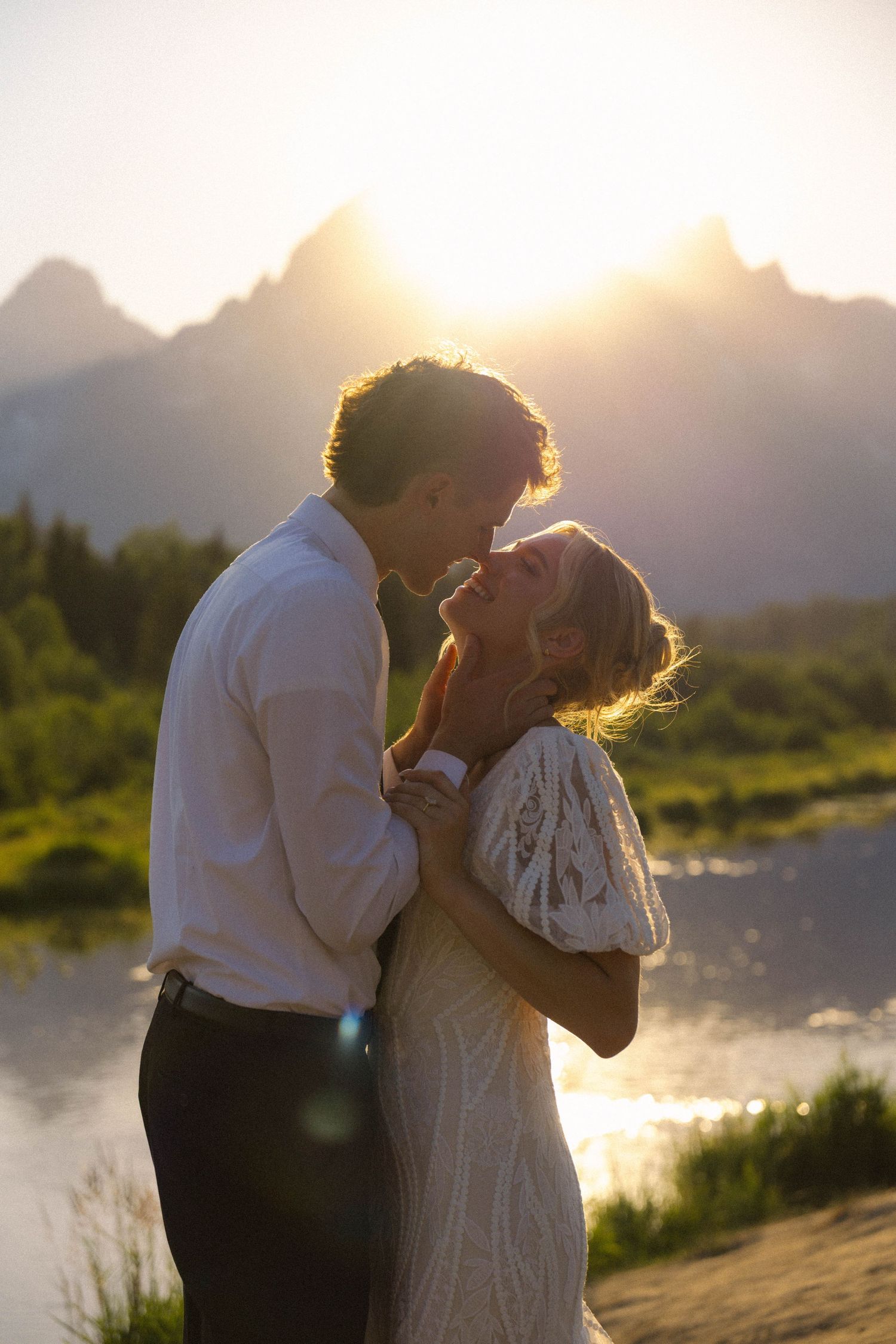 Couple sharing a romantic kiss at golden sunset with mountain lake backdrop, bride in lace dress.