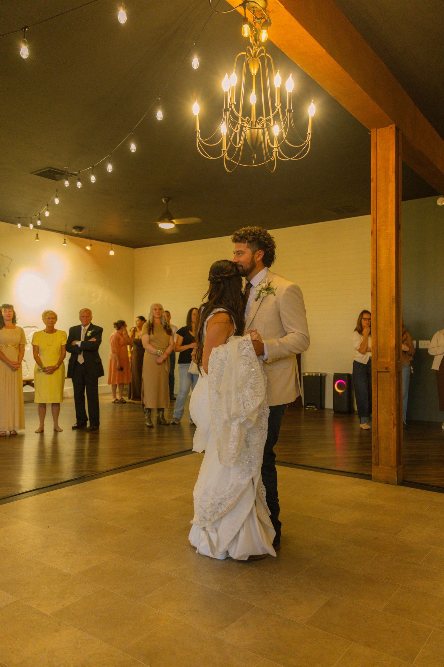 Bride and groom share their first dance under a chandelier while wedding guests watch in a warmly lit reception hall.