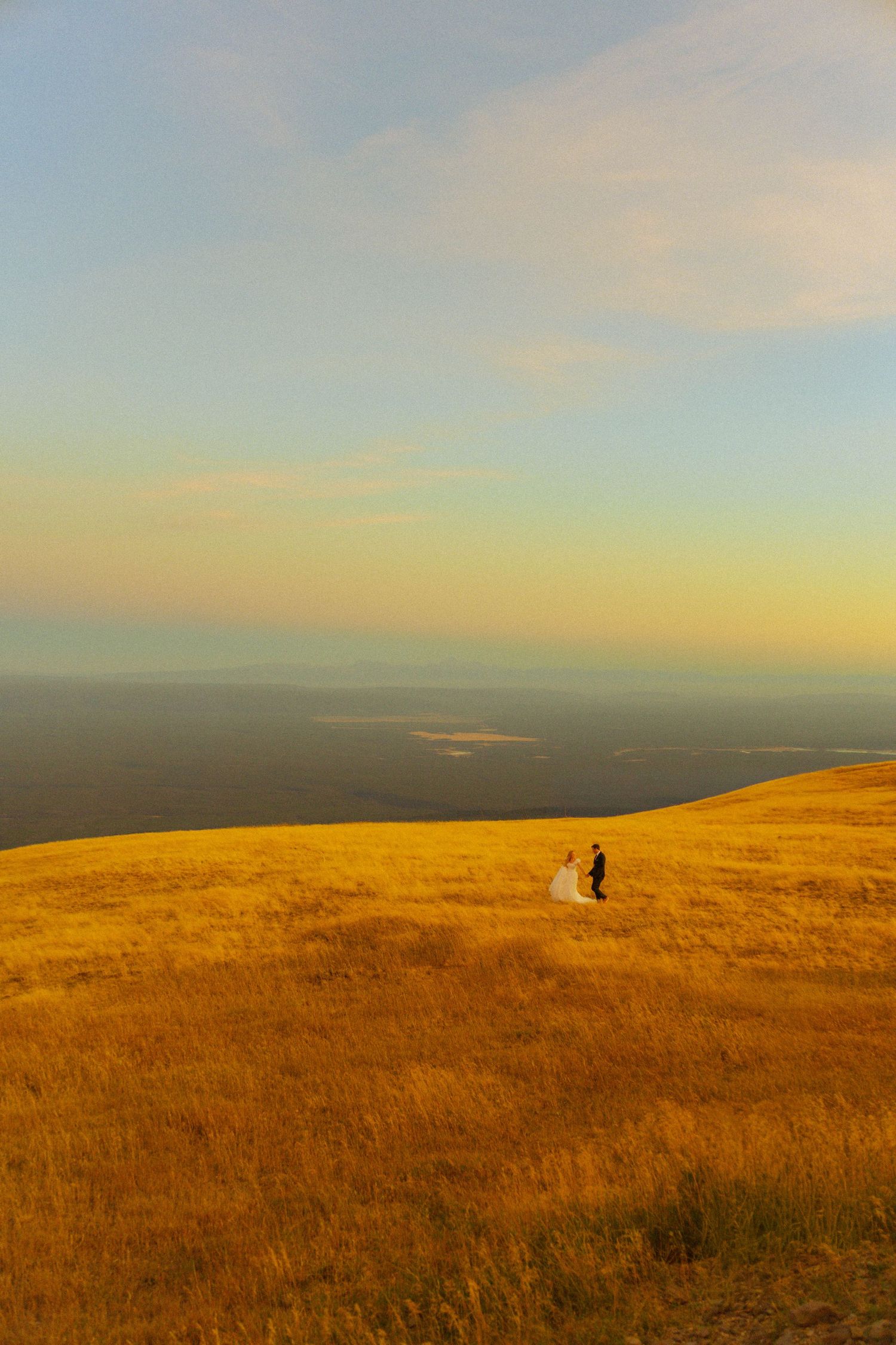 Bride and groom standing on a golden hillside at sunset with a vast landscape stretching into the horizon.