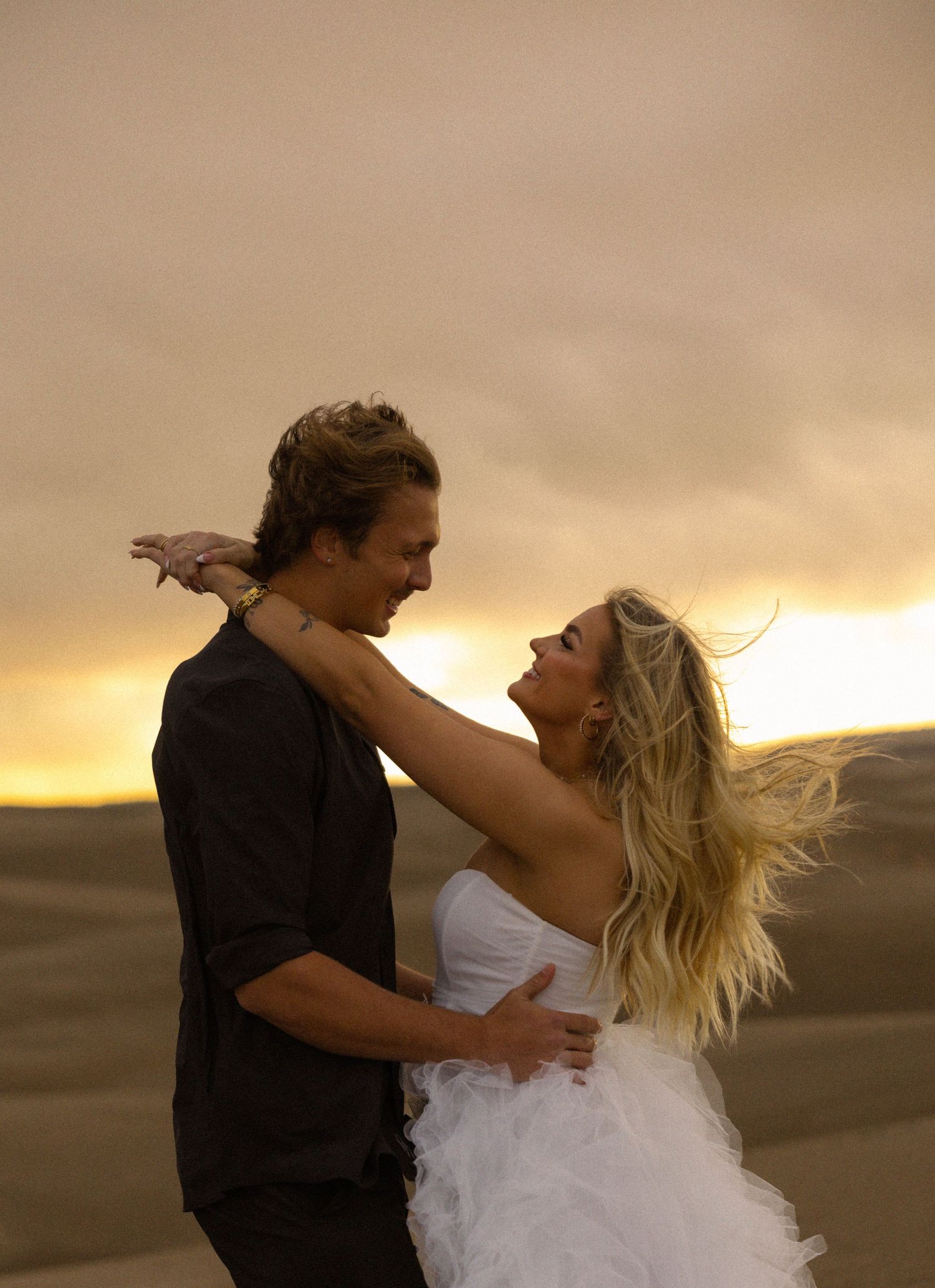 Couple embracing and laughing at sunset in sandy desert dunes, woman in white dress with wind-blown blonde hair.