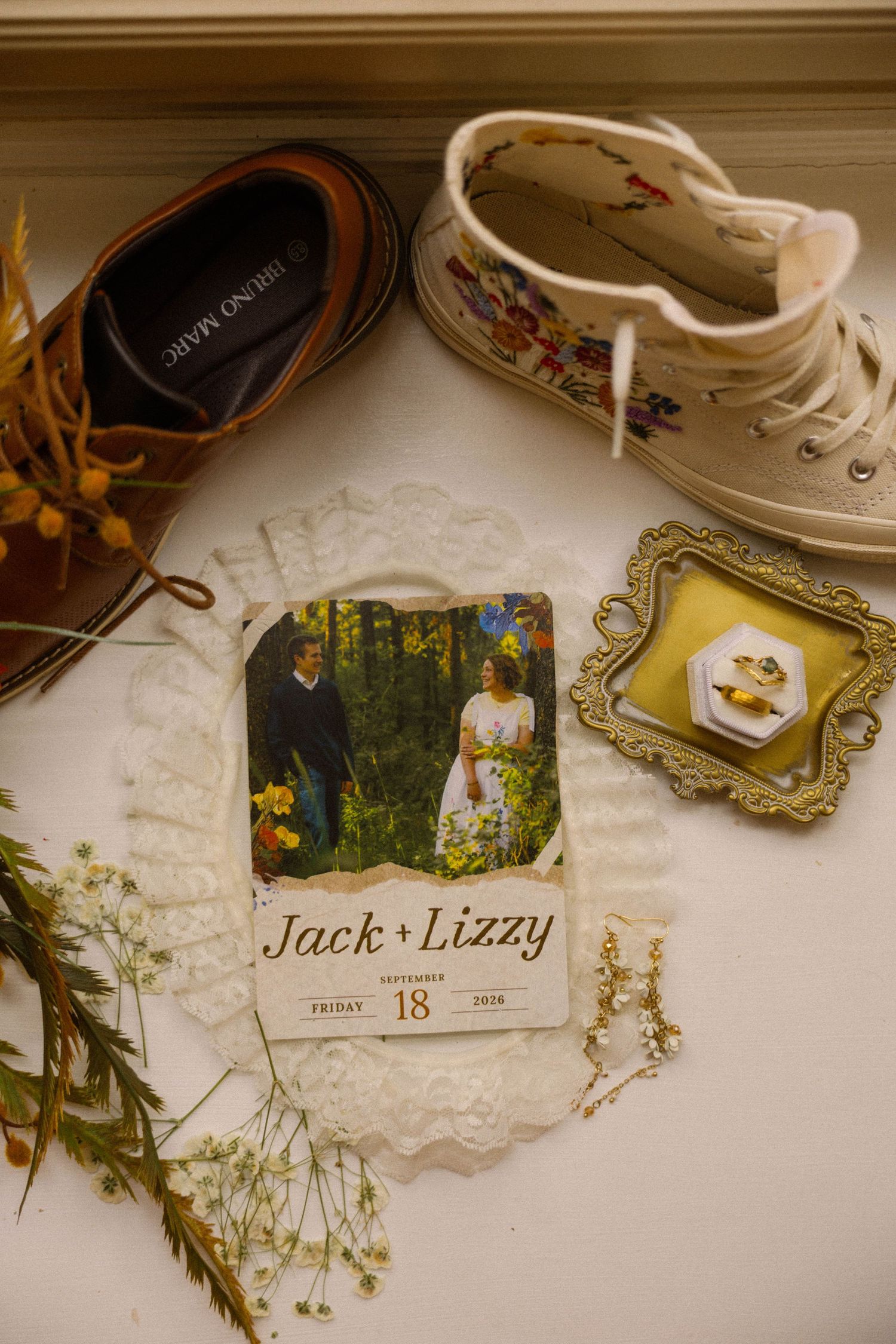Wedding flat lay featuring Jack and Lizzy save-the-date card, floral sneakers, brown dress shoes, gold ring box, and wildflowers.