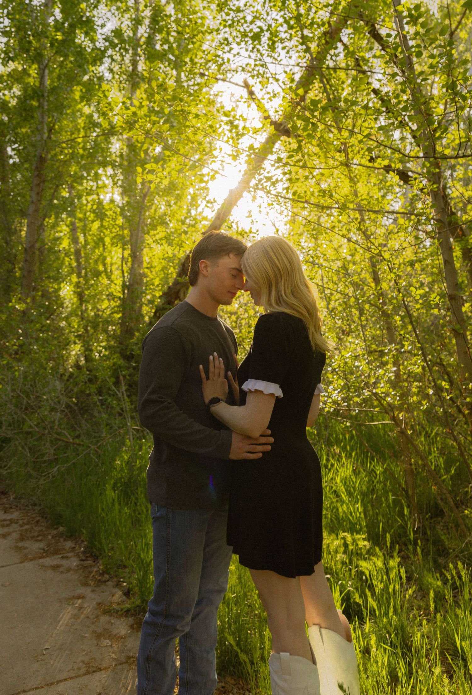 A couple shares a romantic moment on a sunlit forest path surrounded by lush green trees.