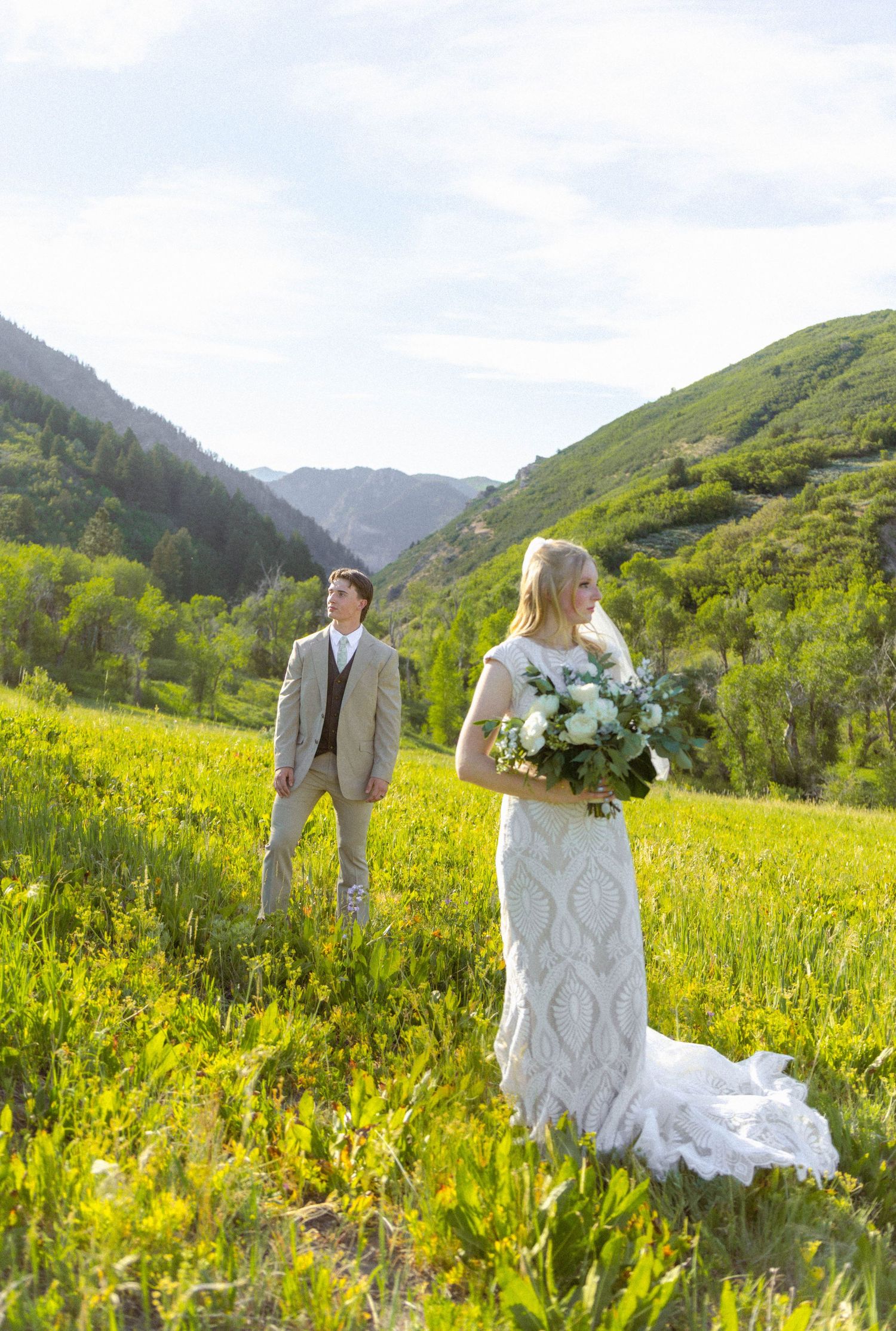 Bride and groom stand in a lush green mountain meadow, bride holding a white bouquet in a lace wedding gown.