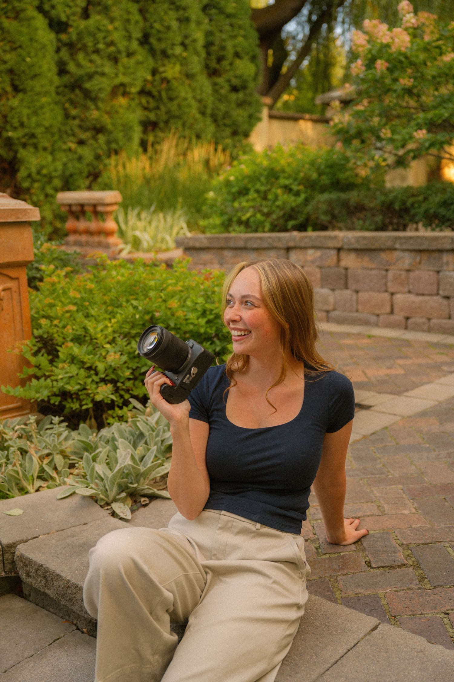 Smiling young woman sitting outdoors holding a DSLR camera in a lush garden setting.
