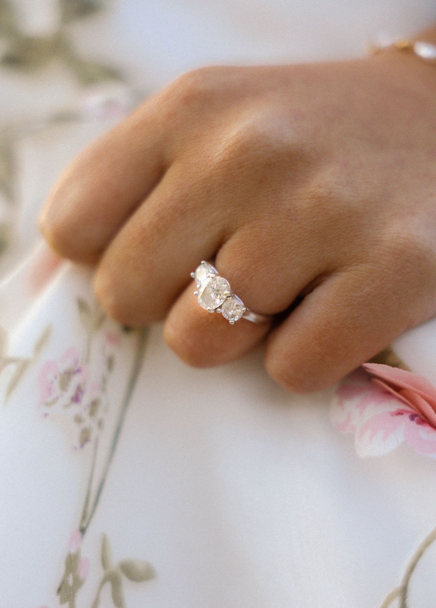 Close-up of a three-stone diamond engagement ring on a bride's hand resting on floral fabric.