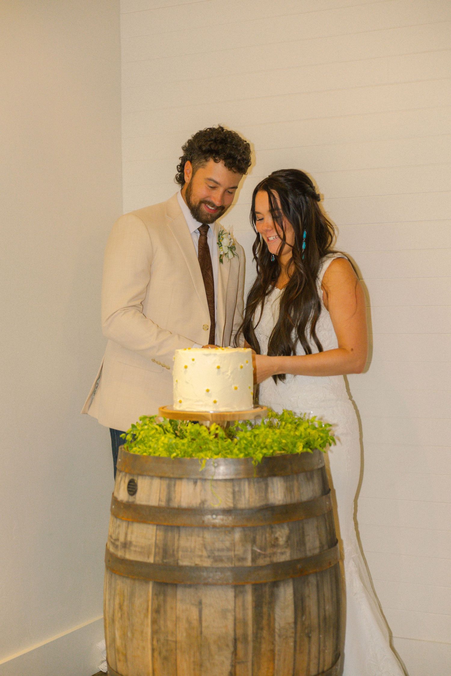Bride and groom cutting a white wedding cake displayed on a rustic wooden barrel at their wedding reception.