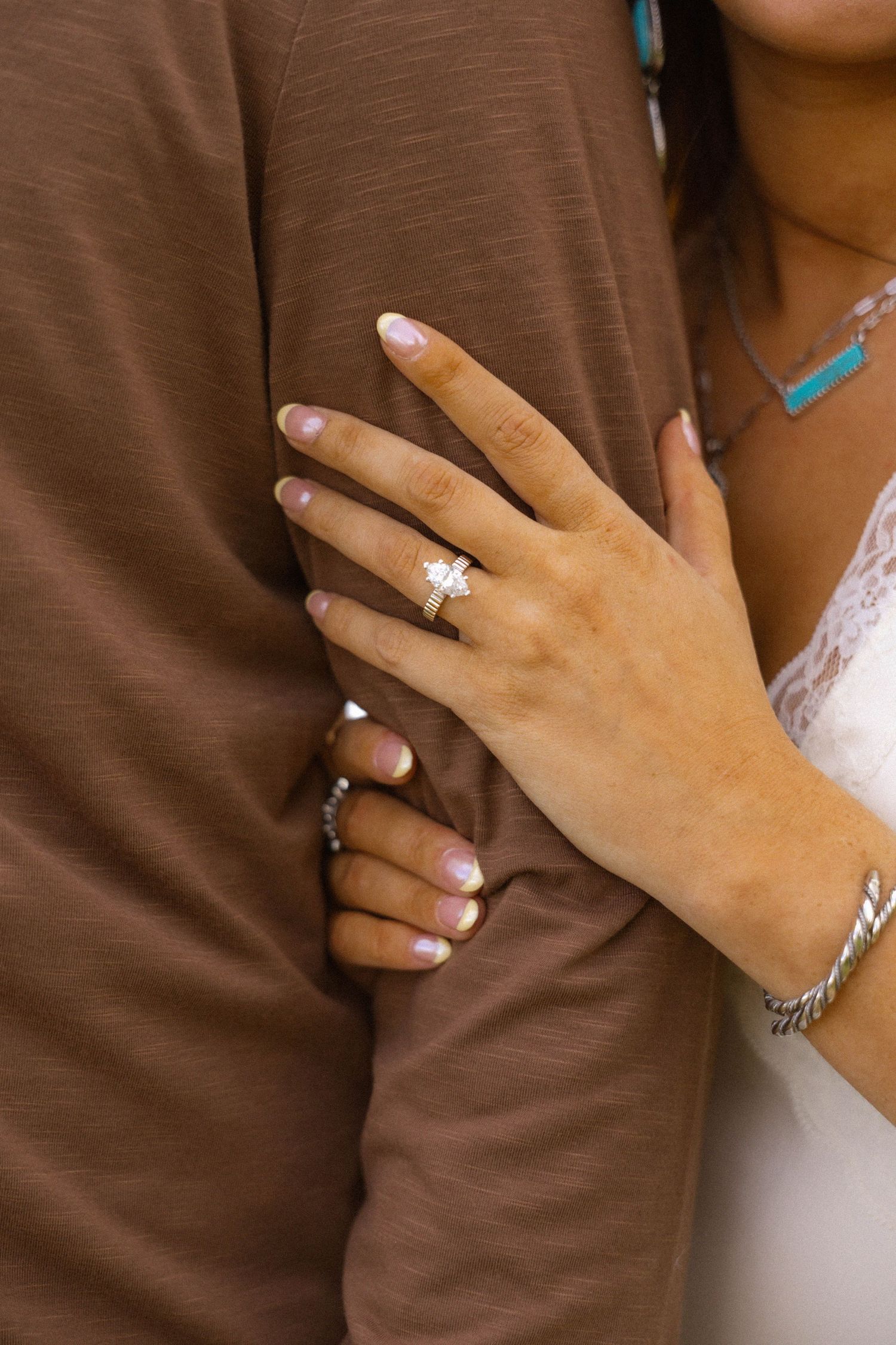 Close-up of a woman's hand displaying a diamond engagement ring while embracing a man in a brown shirt.