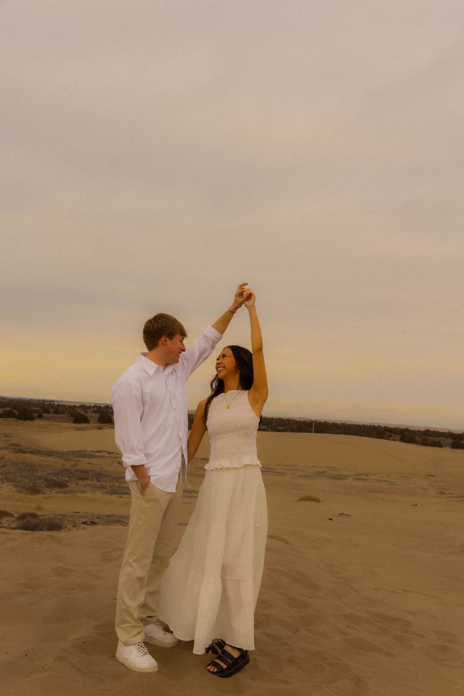 Couple dancing on sandy beach at sunset, woman in white dress spinning, man in white shirt and khakis, romantic golden hour.