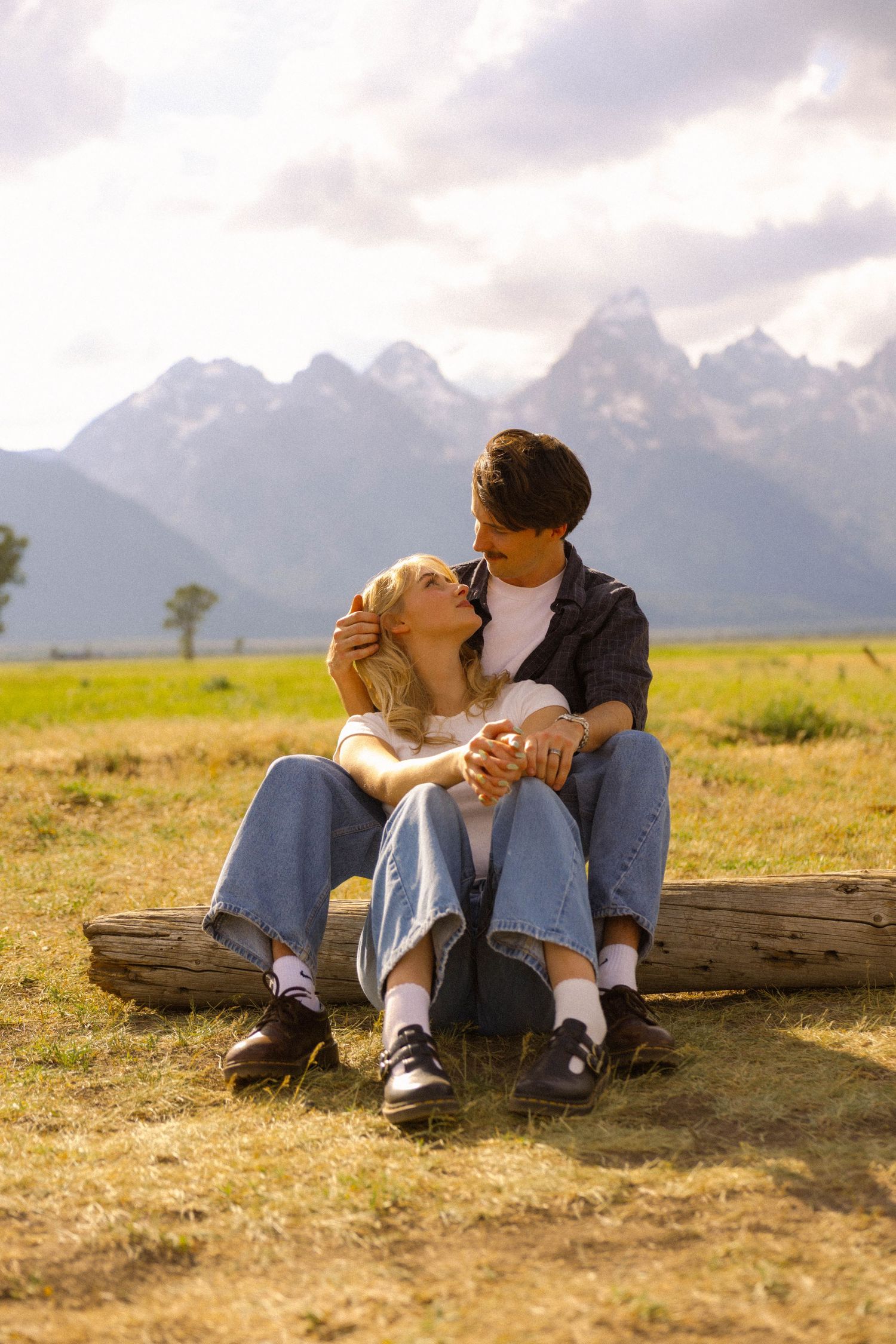 A couple sits on a log in a grassy field, holding hands with the Grand Teton mountains in the background.