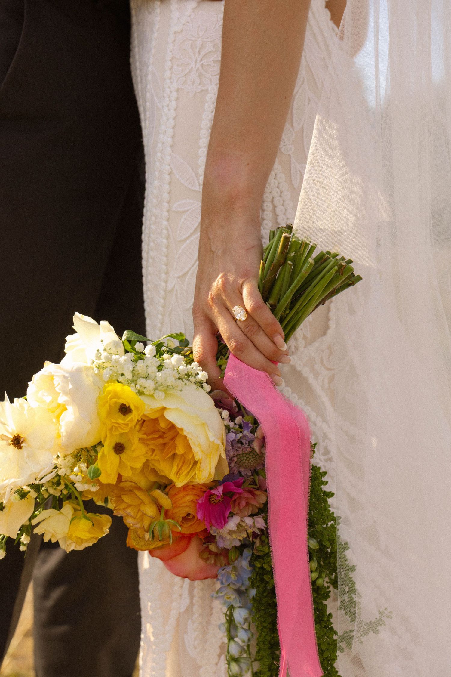 Bride holding colorful wedding bouquet with pink ribbon, wearing white lace dress and gold ring.