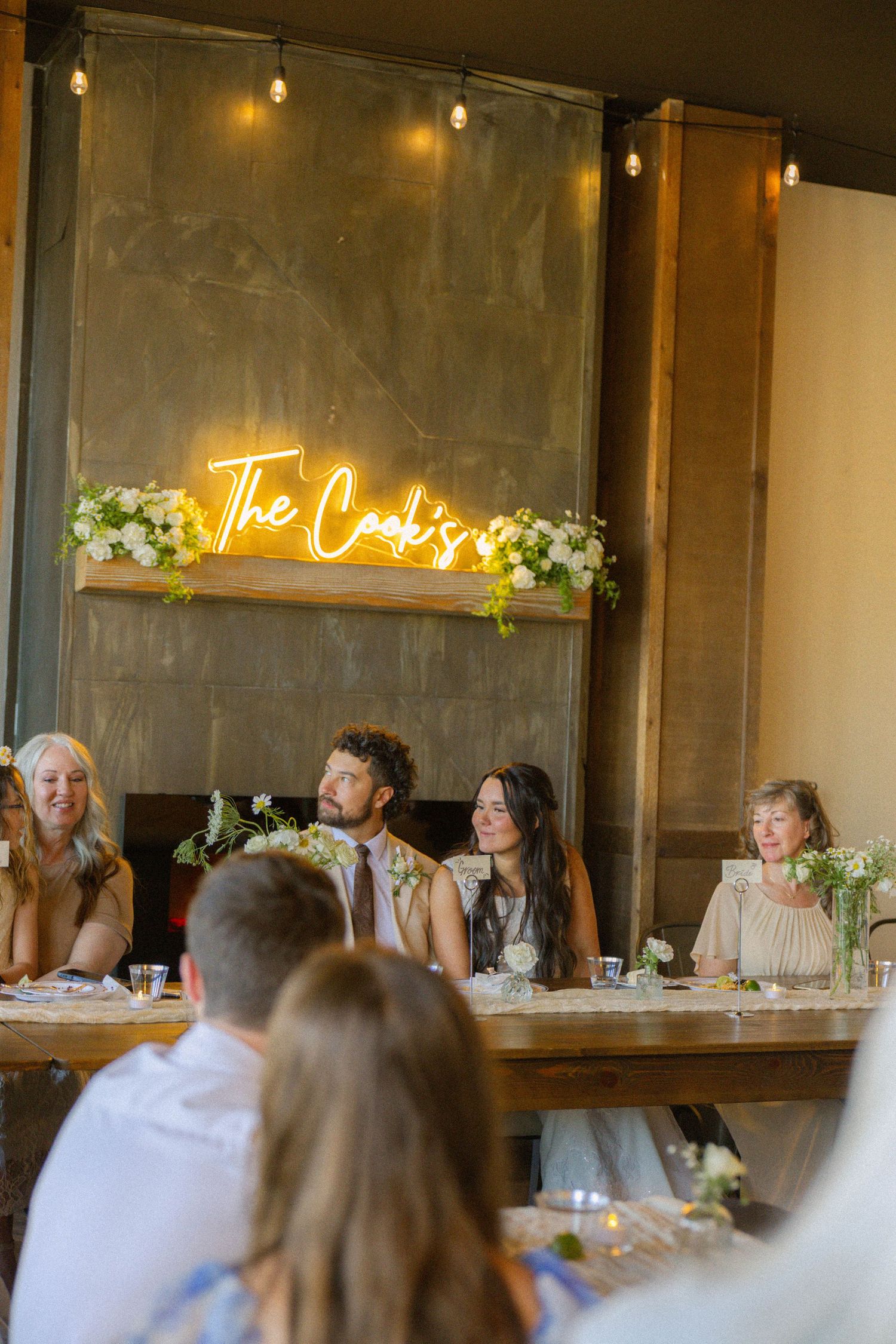 Wedding reception with couple at head table beneath glowing neon sign reading The Cooks, decorated with white floral arrangements.