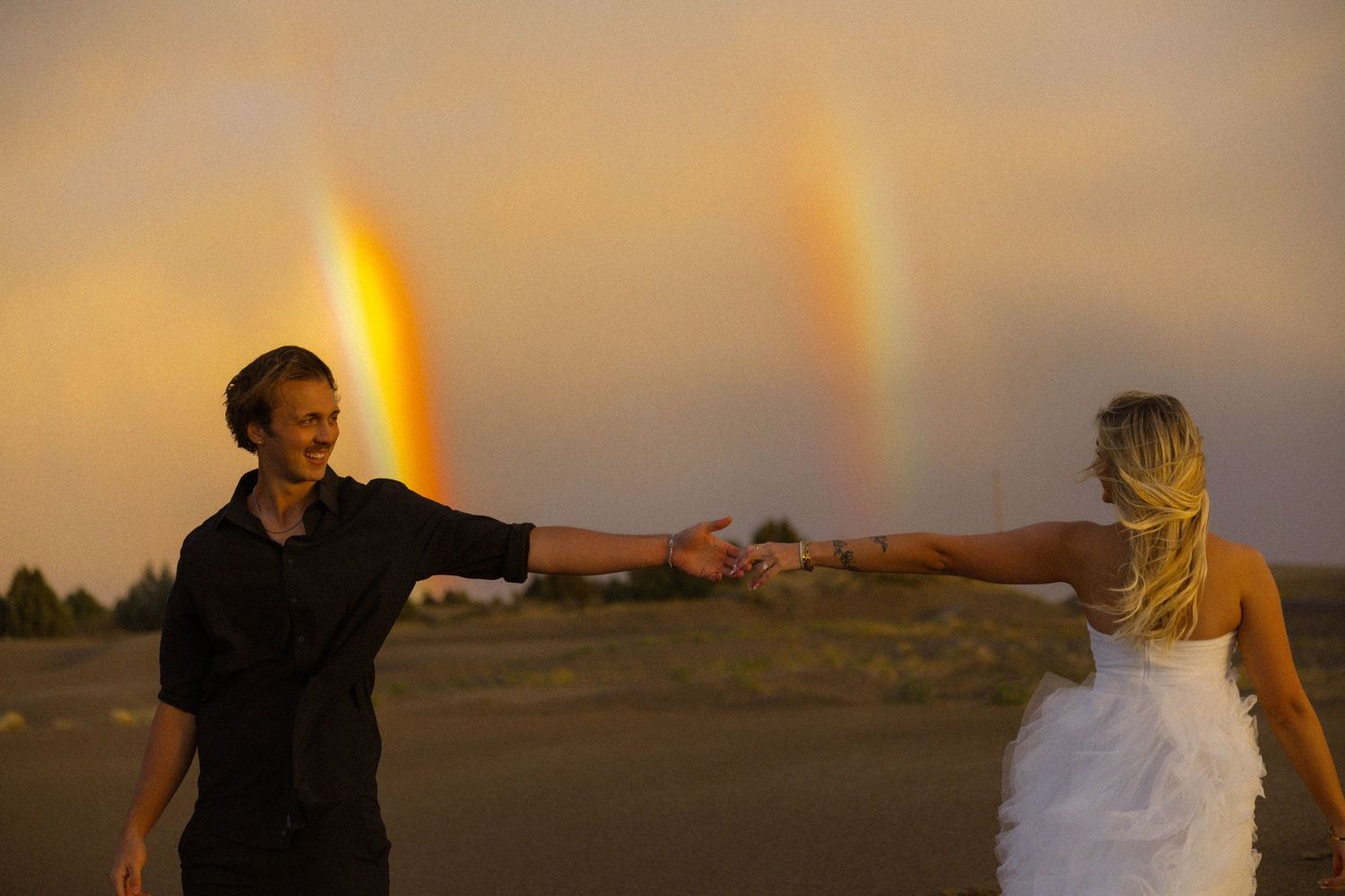 Bride and groom hold hands at sunset with a double rainbow glowing in the golden sky behind them.