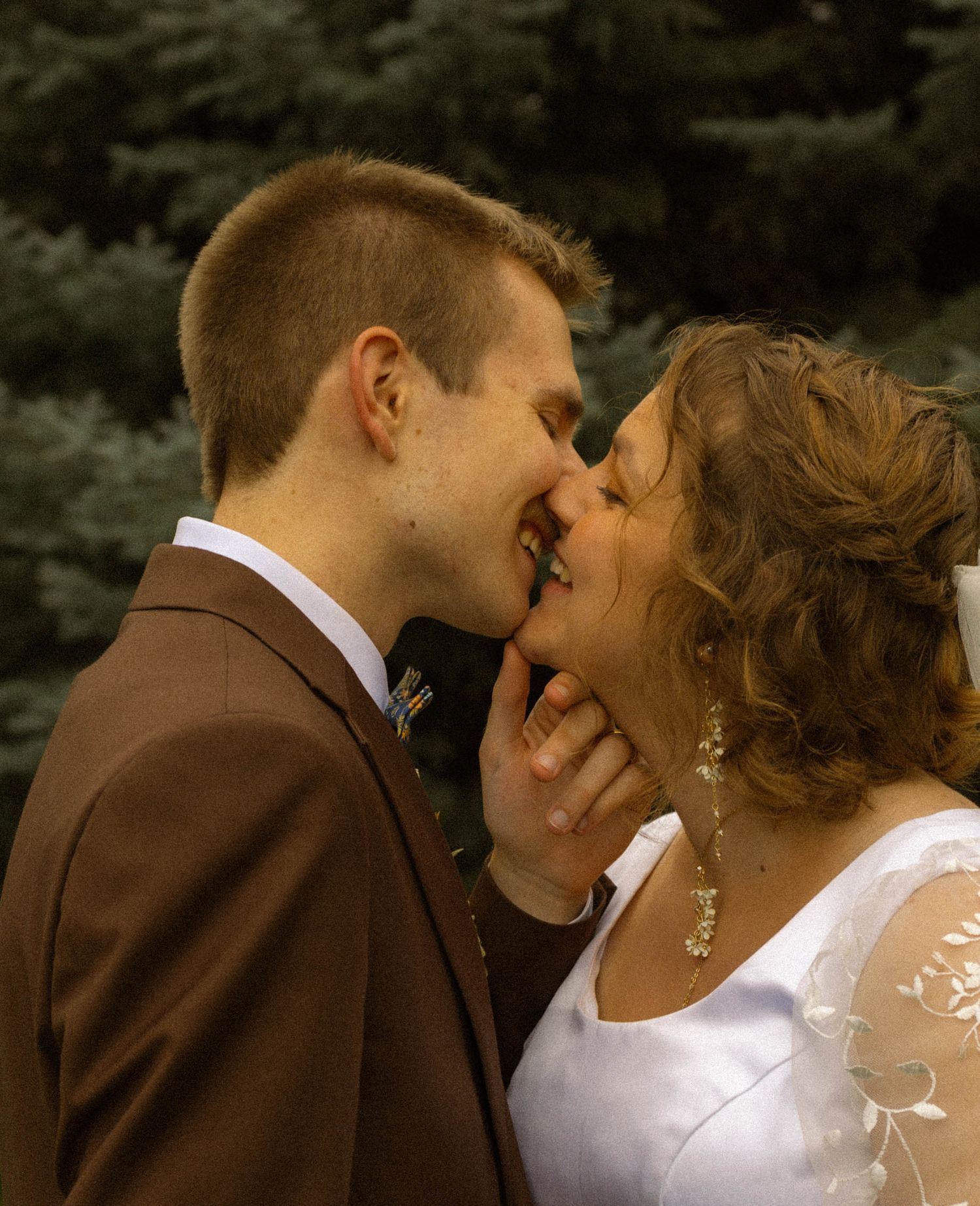Bride and groom sharing a romantic kiss outdoors, with the bride in white and groom in a brown suit.