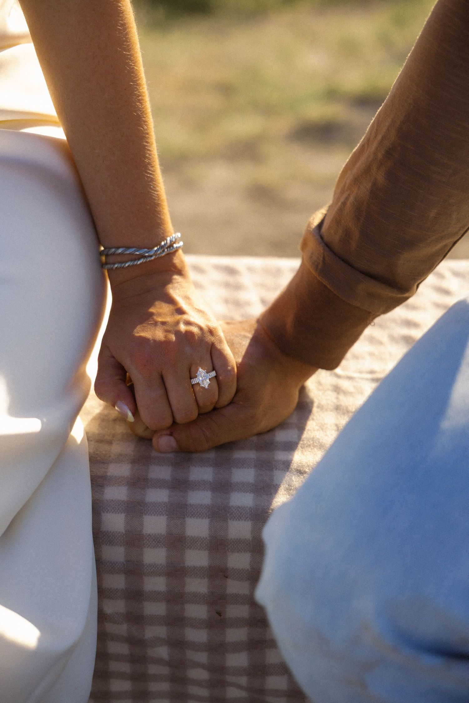 Close-up of couple holding hands with diamond engagement ring and silver bracelet on checkered blanket.