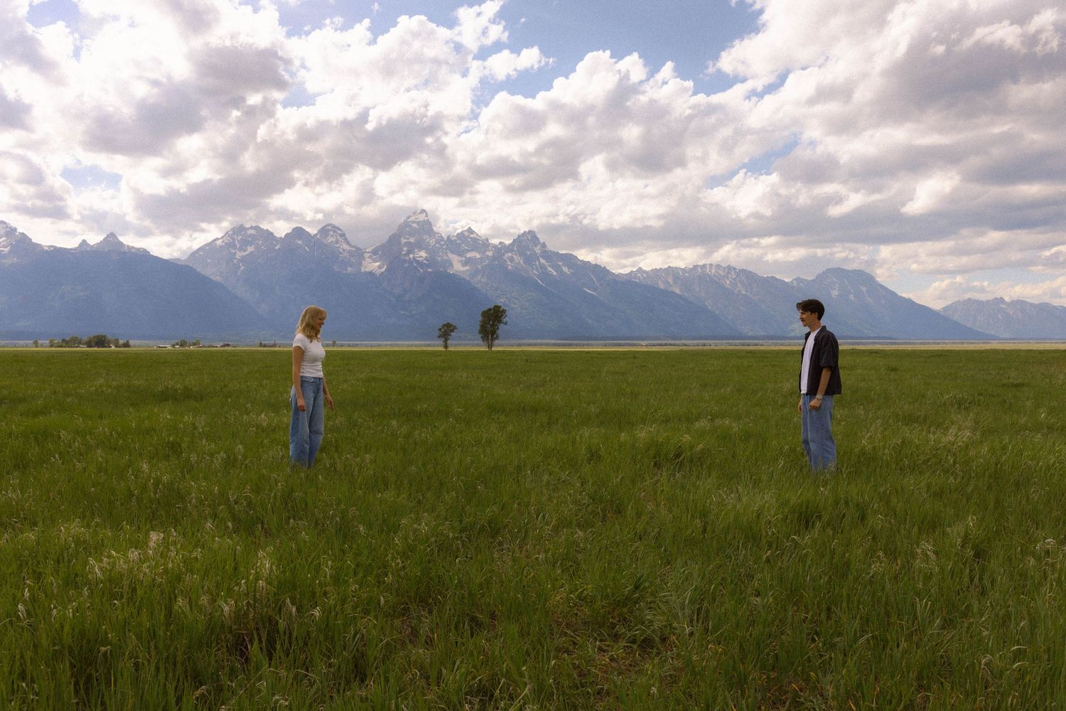Two people stand apart in a vast green meadow with the Grand Teton mountain range under a cloudy sky.